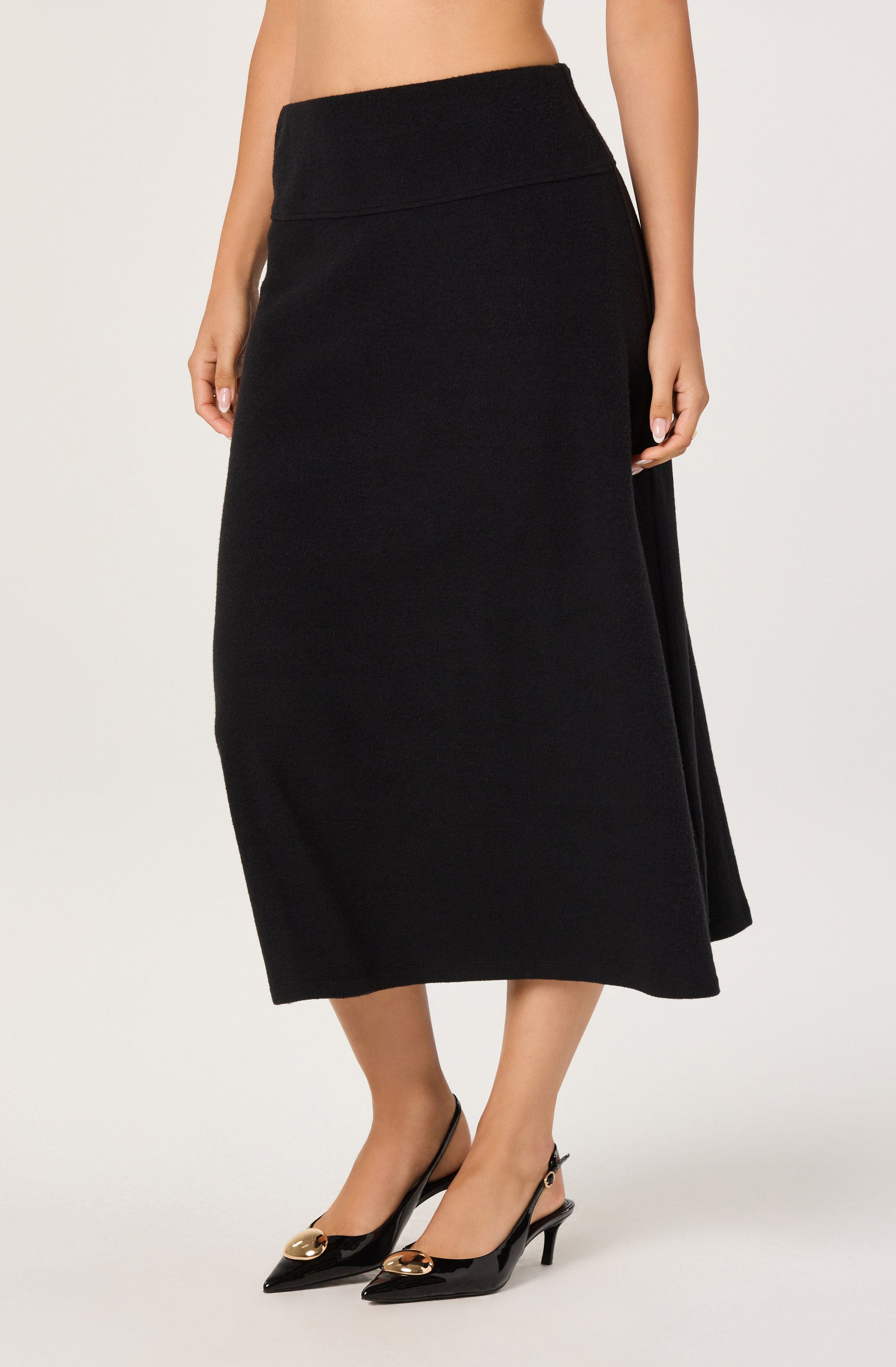 A woman is wearing a long, high-waisted black skirt that extends to mid-calf length. The skirt appears to be made of a textured fabric and has a fitted waistband. She is also wearing black pointed-toe high heels with gold embellishments on the front straps.