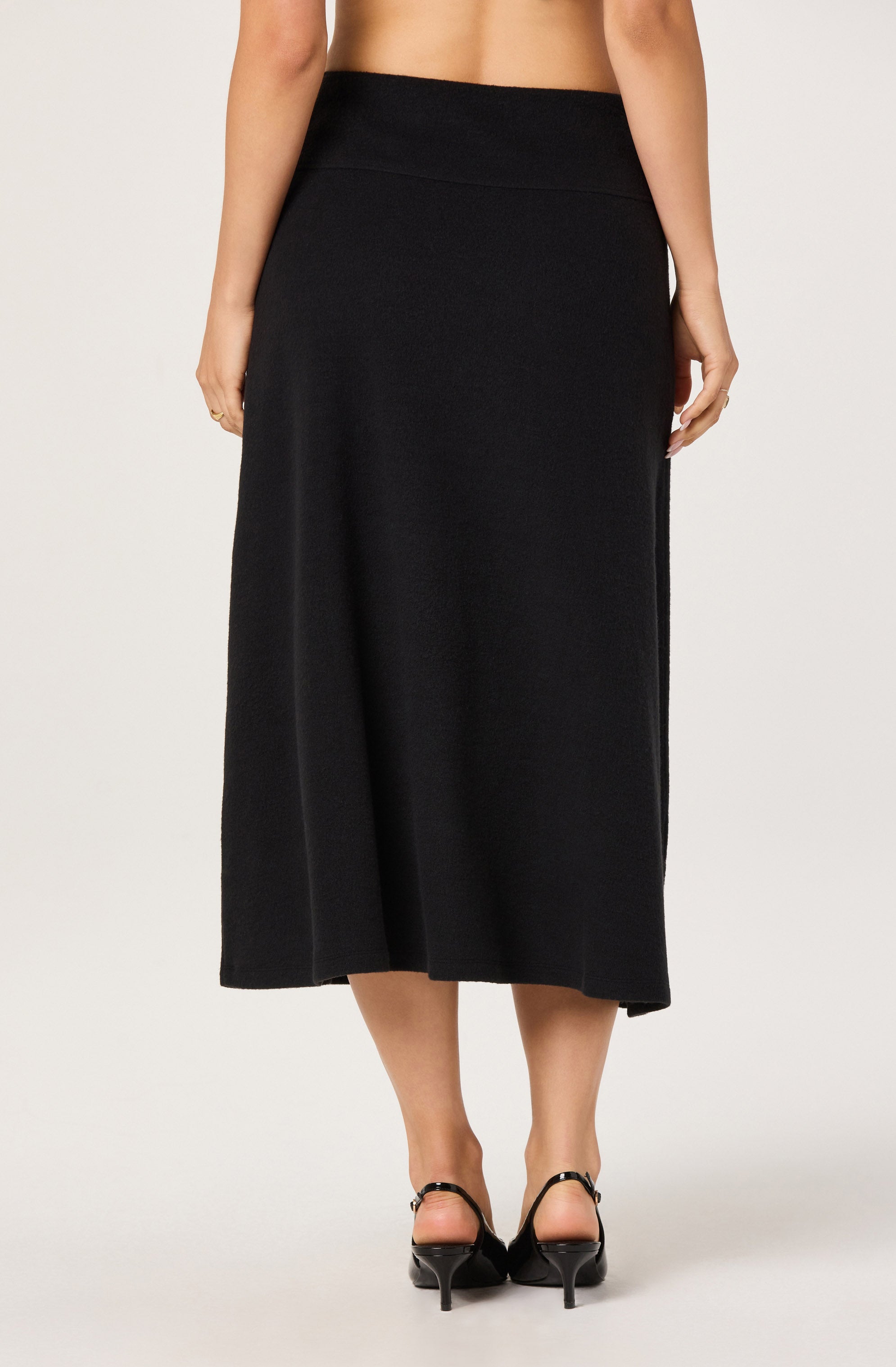 A woman is wearing a high-waisted, ankle-length black skirt with a simple, smooth appearance. She is also wearing black high-heeled shoes with ankle straps.