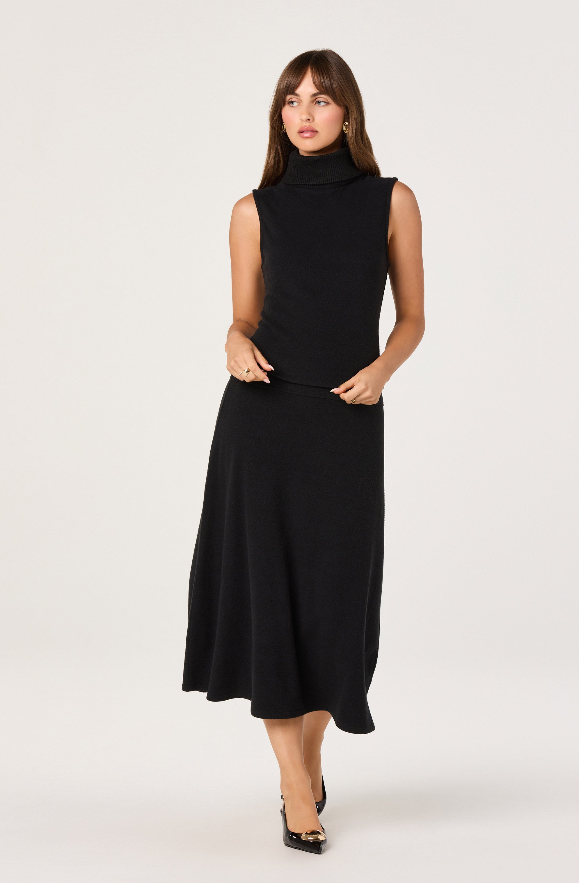 A woman is wearing a sleeveless, black turtleneck top made of a ribbed knit material. She pairs it with a black, ankle-length, A-line skirt that drapes softly down to her ankles. She is also wearing black pointed-toe heels with a gold accent on the toe box.