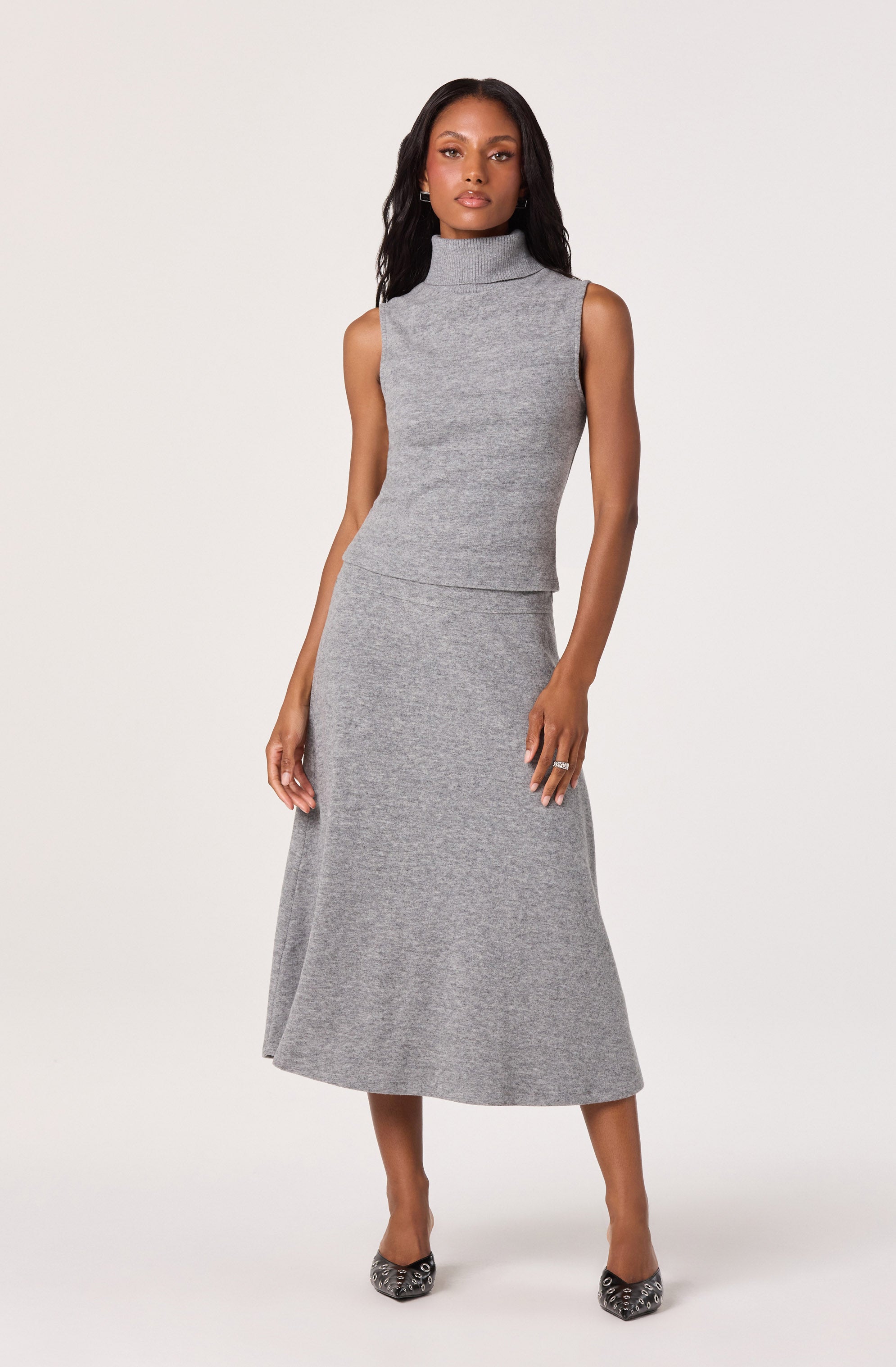 A woman is wearing a sleeveless, high-neck, knit top and a matching knit midi skirt, both in a light gray color. She pairs the outfit with pointed-toe shoes that have a patterned design.