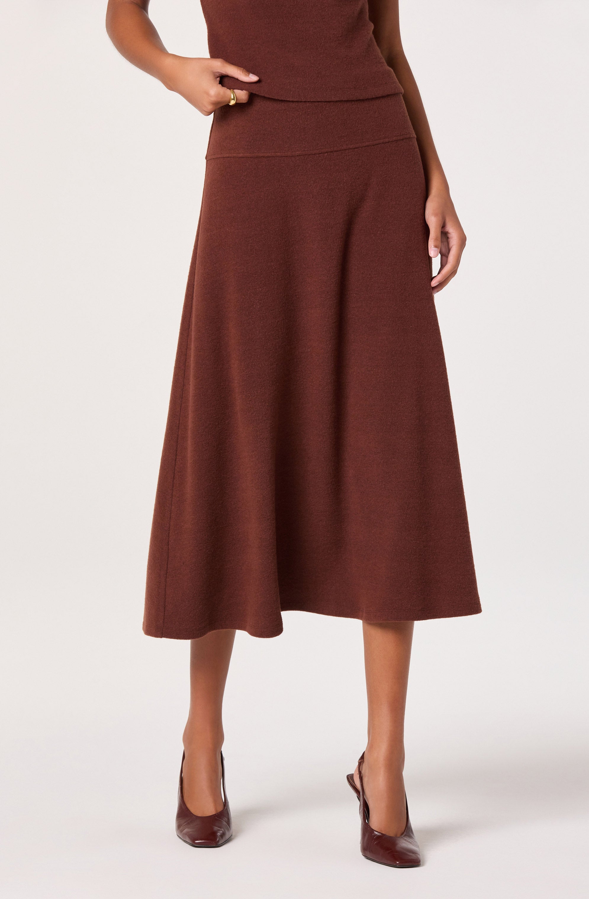 A woman is wearing a matching burgundy two-piece outfit consisting of a sleeveless top and a high-waisted, A-line midi skirt that falls below the knees. She has paired the outfit with burgundy high-heeled shoes. The fabric of the clothing appears soft and textured.