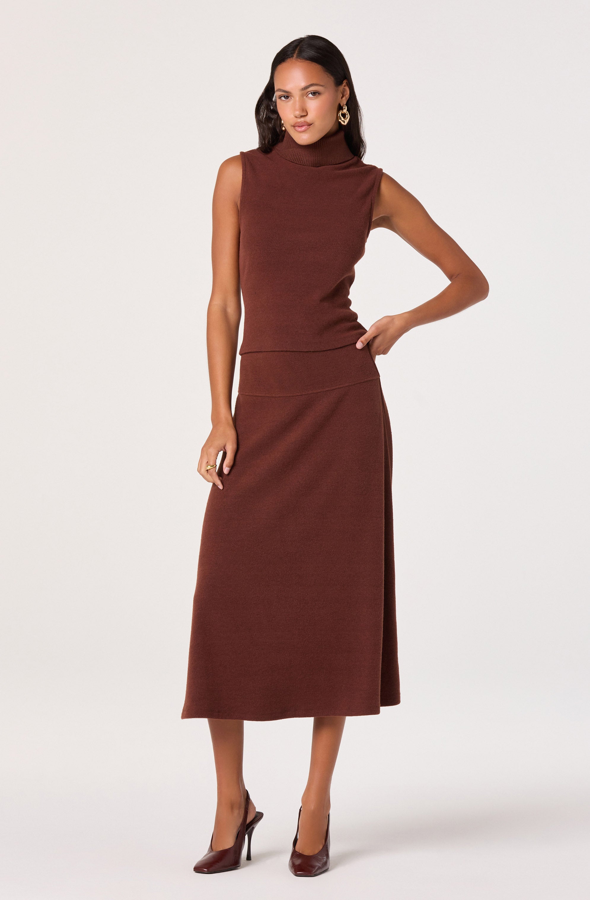 A woman is wearing a sleeveless, high-neck, fitted maroon top paired with a long, matching maroon skirt that reaches her calves. She is also wearing matching maroon high heels.