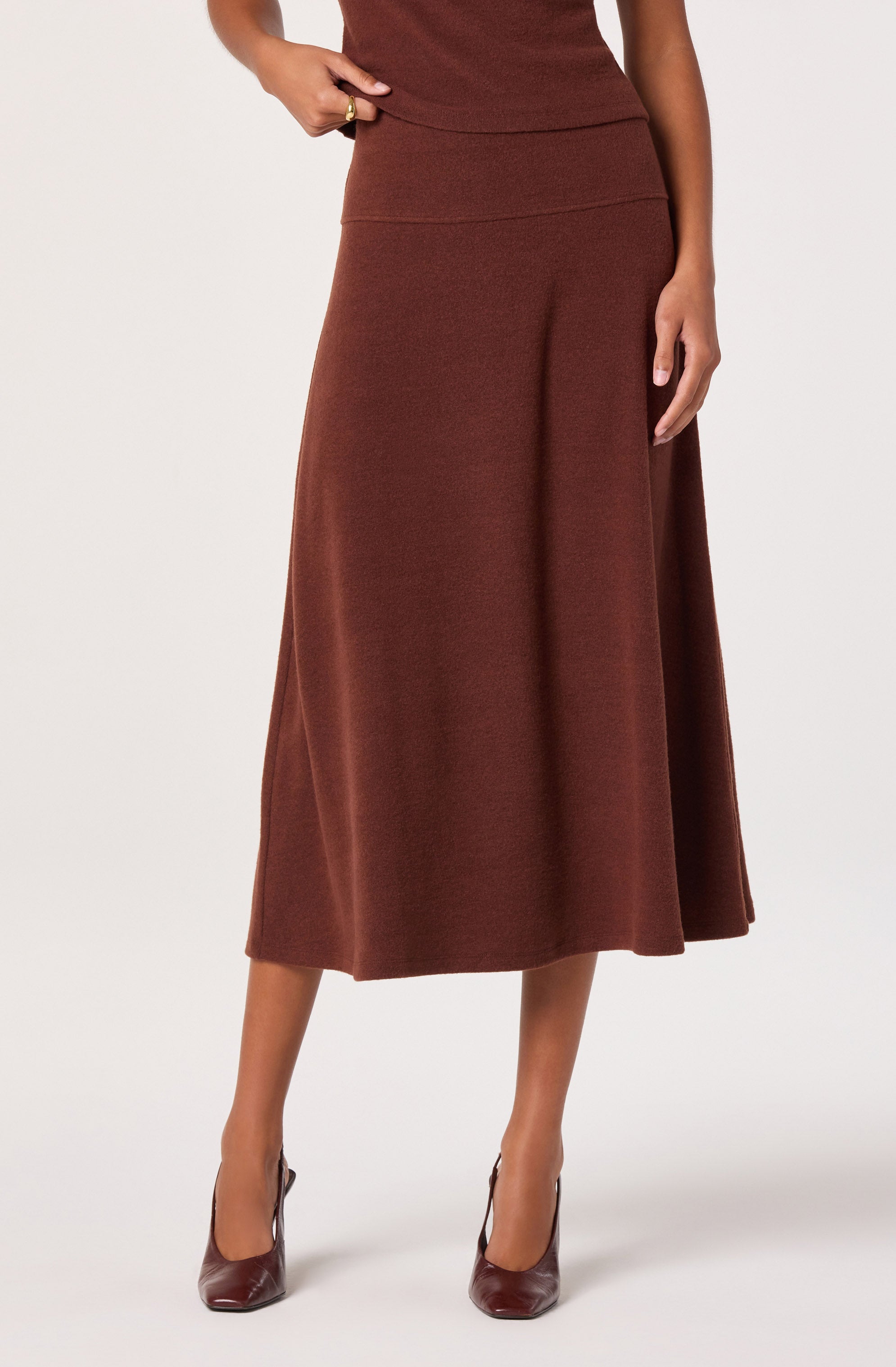 A woman is wearing a long, high-waisted, A-line skirt in a rich brown color with a textured fabric. She pairs it with matching brown high-heeled shoes and a sleeveless brown top. She has one hand on her hip and the other resting at her side.