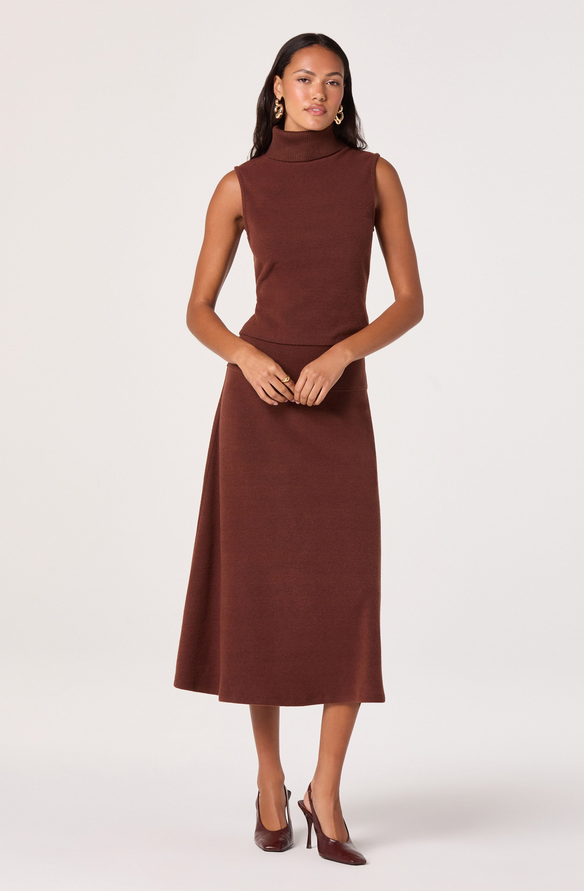 A woman is wearing a sleeveless, high-neck, brown knit top paired with a matching long, straight skirt that reaches mid-calf. She is accessorized with gold hoop earrings and a gold bracelet, and is wearing brown high-heeled shoes.