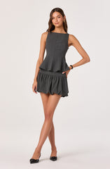 A woman is wearing a sleeveless, dark gray, tailored top with vertical seam details and a peplum hem. She pairs it with matching dark gray, ruffled shorts that have an elastic waistband and a loose, gathered fit. She completes her outfit with black pointed-toe heels and gold hoop earrings.