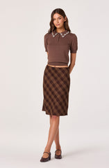 A woman is wearing a short-sleeved, brown knit top with a decorative collar adorned with beads or embroidery. She pairs it with a high-waisted, knee-length plaid skirt in shades of brown and yellow. She completes the outfit with dark brown, closed-toe heels.