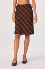 A woman is wearing a knee-length skirt with a brown plaid pattern, featuring intersecting yellow and dark brown lines. The skirt has a small bow detail at the waistband. She is also wearing brown heeled shoes with ankle straps.