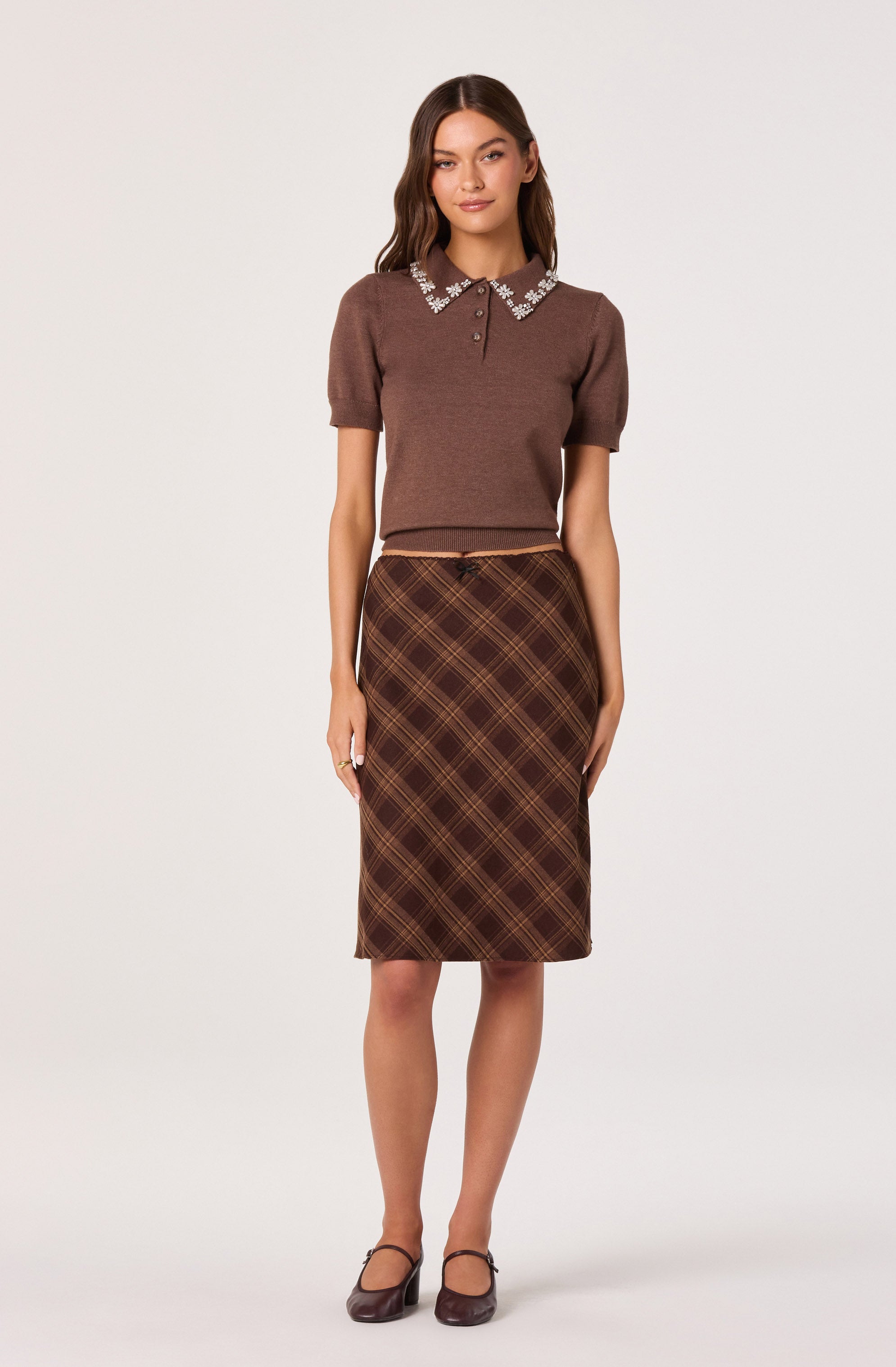 The woman is wearing a short-sleeved, brown knit top with a collar decorated with white pearls and small floral embellishments. She pairs it with a high-waisted, knee-length brown plaid skirt featuring a plaid pattern in shades of brown and yellow. She completes the outfit with brown heels.