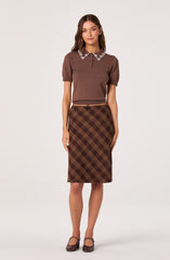 The woman is wearing a short-sleeved, brown knit top with a collar decorated with white pearls and small floral embellishments. She pairs it with a high-waisted, knee-length brown plaid skirt featuring a plaid pattern in shades of brown and yellow. She completes the outfit with brown heels.
