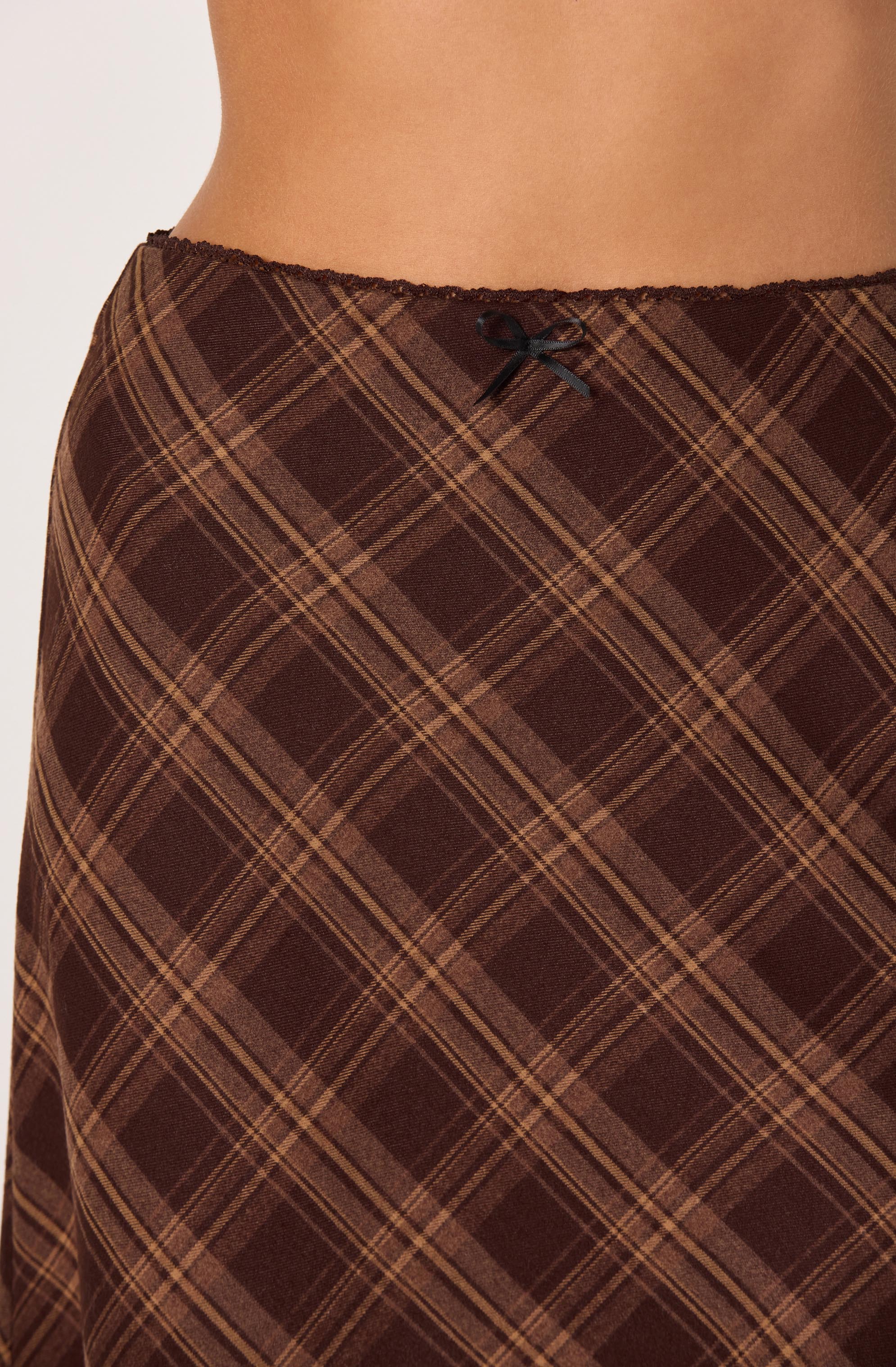 A close-up of a person's waist and hips, showing a brown plaid skirt with orange and beige lines in a diagonal pattern. The skirt has a small black bow at the waistband. The waistband appears soft and has a black lace trim. The fabric of the skirt looks smooth and slightly shiny.