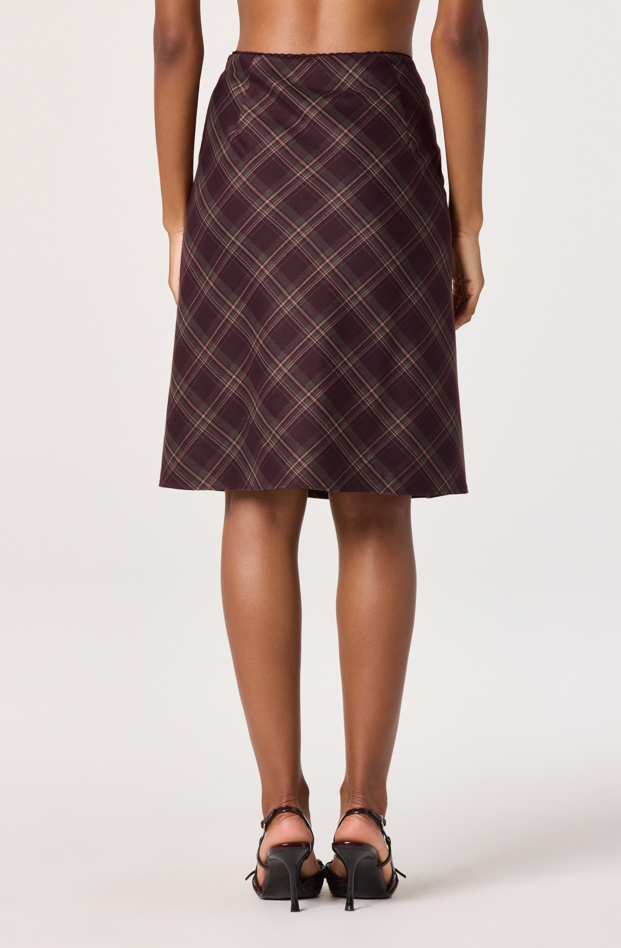 A woman is wearing a high-waisted, knee-length skirt with a plaid pattern in shades of dark red, black, and beige. She is also wearing black high-heeled shoes with ankle straps.