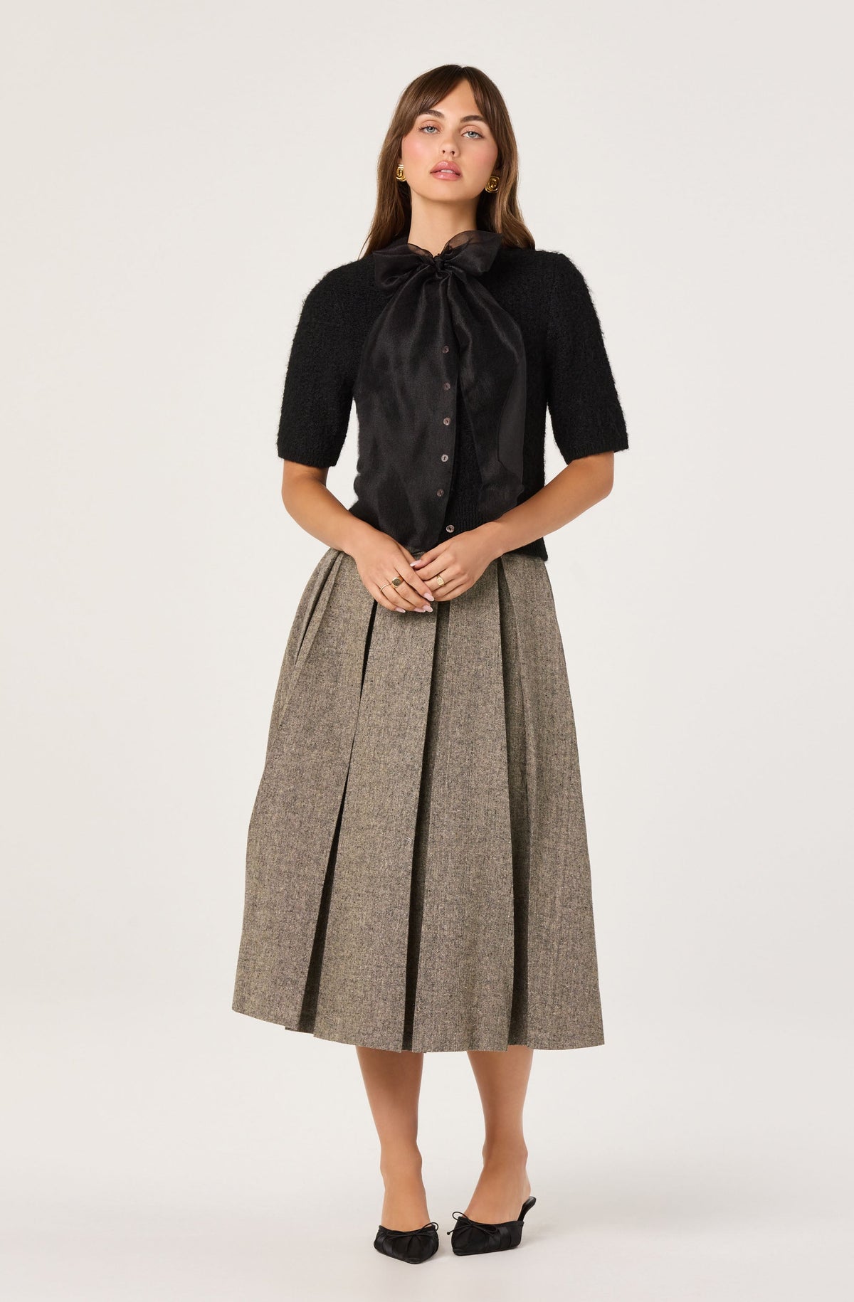 A woman is wearing a black blouse with a large, satin bow at the collar, and a pair of high-waisted, pleated, ankle-length skirt in a brown tweed pattern with darker vertical lines. She is also wearing black heeled shoes with bow accents.