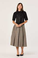 A woman is wearing a black blouse with a large, satin bow at the collar, and a pair of high-waisted, pleated, ankle-length skirt in a brown tweed pattern with darker vertical lines. She is also wearing black heeled shoes with bow accents.