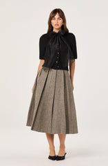 A woman is wearing a black short-sleeved top with a large satin bow at the neckline and small buttons down the front. She pairs it with a high-waisted, pleated, midi-length skirt made of a beige and brown checkered fabric. She is also wearing black pointed-toe heels with a bow detail.