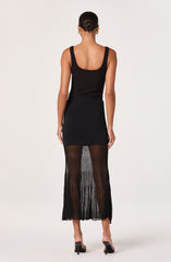 A woman is wearing a sleeveless black top with thick straps and a scoop neckline. She has paired it with a black skirt that is fitted at the waist and made of a semi-sheer, pleated fabric that extends to her ankles. She is also wearing black high-heeled sandals.