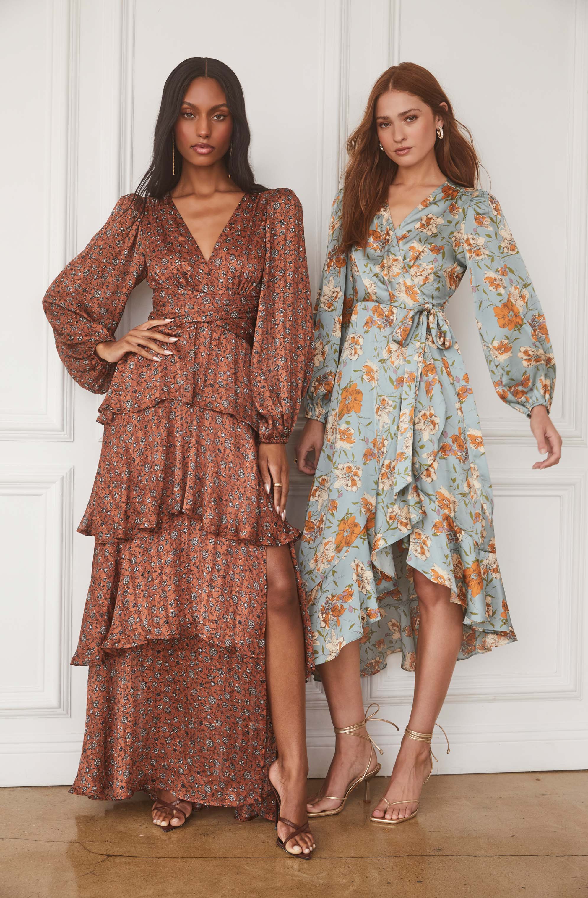 Two women are wearing long, flowy dresses with floral patterns. The woman on the left has a rust-colored dress with small floral prints, long puffed sleeves, a V-neckline, and tiered ruffles. The woman on the right is wearing a light blue dress with orange and white floral designs, a wrap style with a tie at the waist, and a high-low hemline. Both women are wearing open-toed heels, one in dark brown and the other in nude with ankle straps.