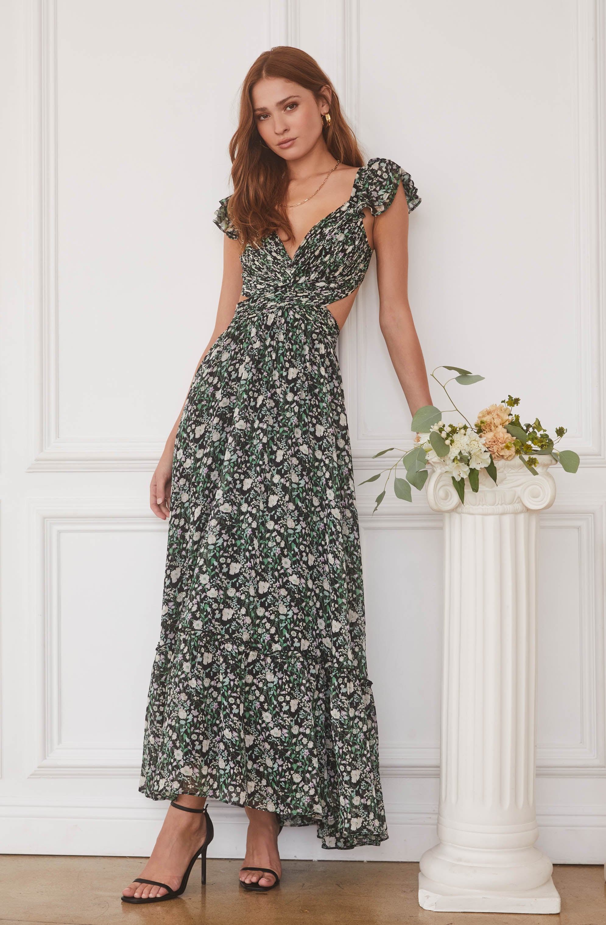 A woman is wearing a long, flowy black dress with a floral pattern in white, purple, and green. The dress features cutouts at the waist and ruffled cap sleeves. She is also wearing black high-heeled sandals.