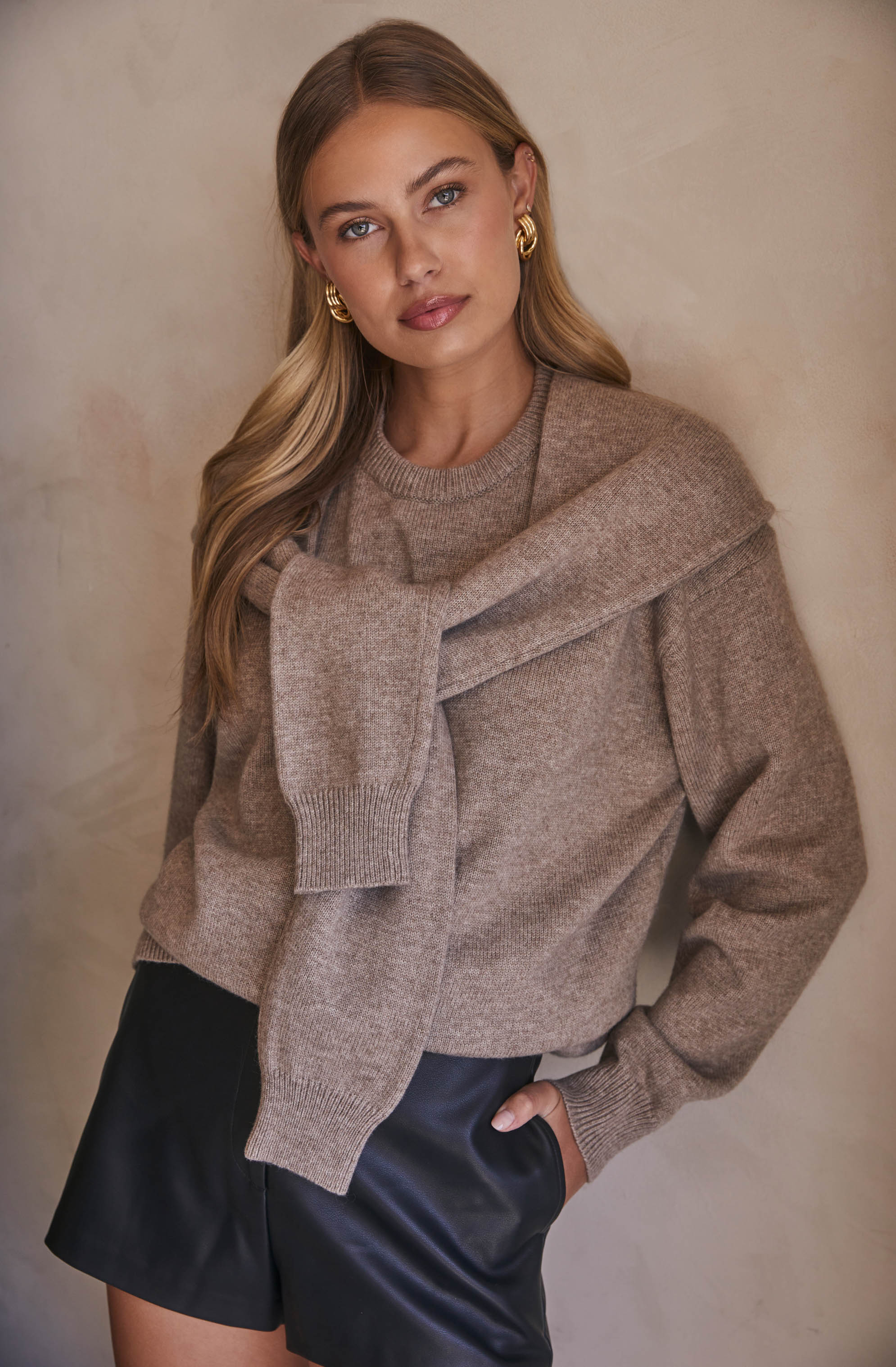 A woman is wearing a beige knit sweater with a crew neckline and long sleeves. She has a matching beige sweater draped over her shoulders. She is also wearing black leather shorts and accessorizing with large gold hoop earrings.