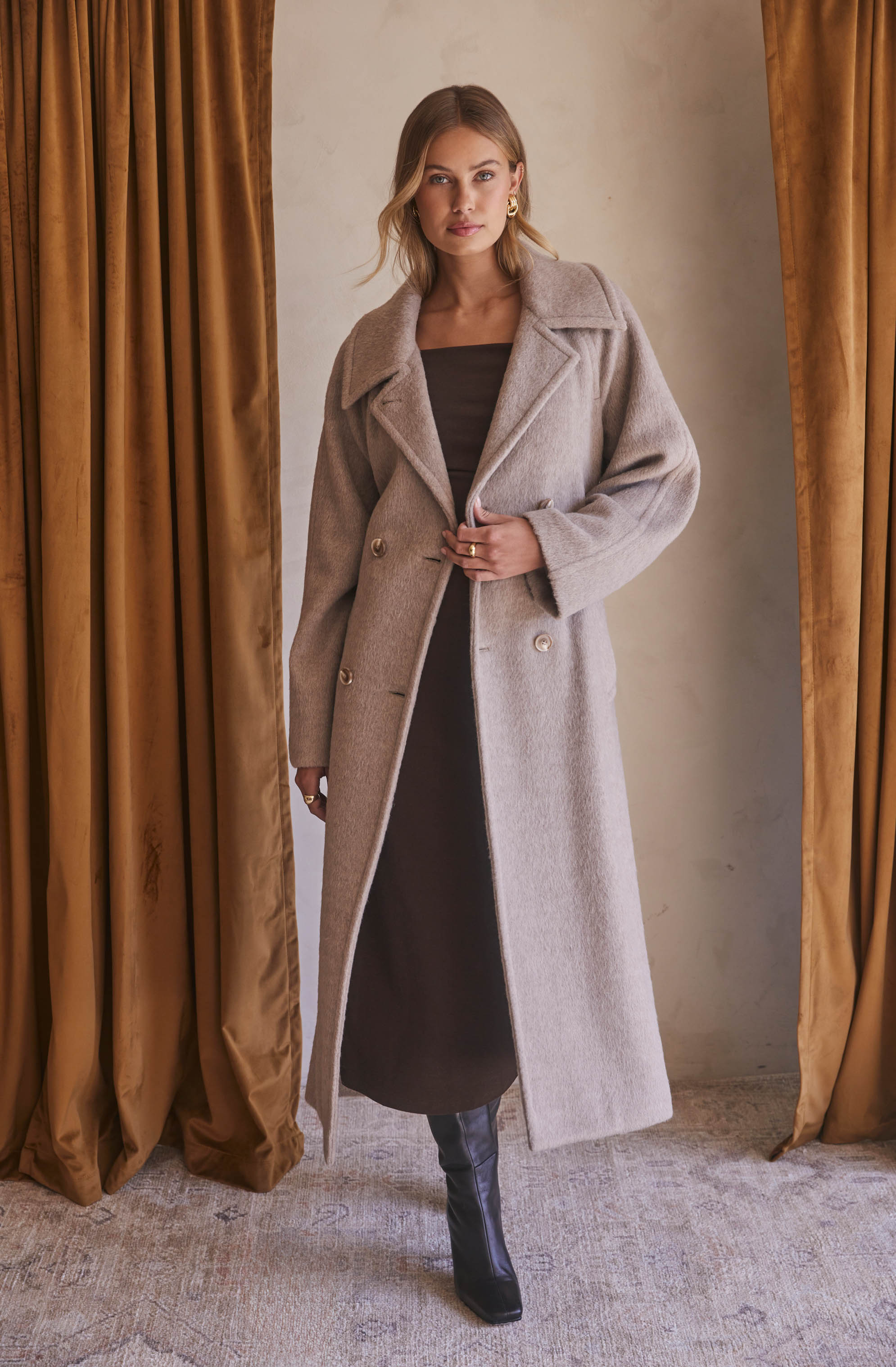 A woman is wearing a long, light beige, double-breasted wool coat with wide lapels. Underneath, she has on a black, ankle-length dress with a high neckline. She is also wearing black, knee-high boots with a slight heel.