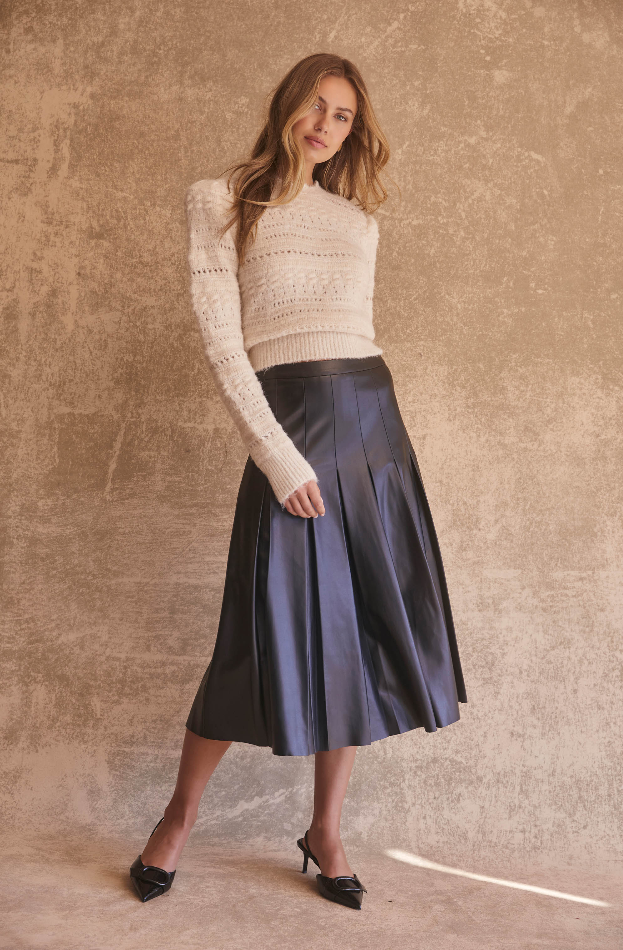 A woman is wearing a cream-colored, knitted sweater with openwork details and long sleeves. She pairs it with a high-waisted, dark blue, pleated midi skirt made of shiny material. She completes the outfit with black pointed-toe heels.
