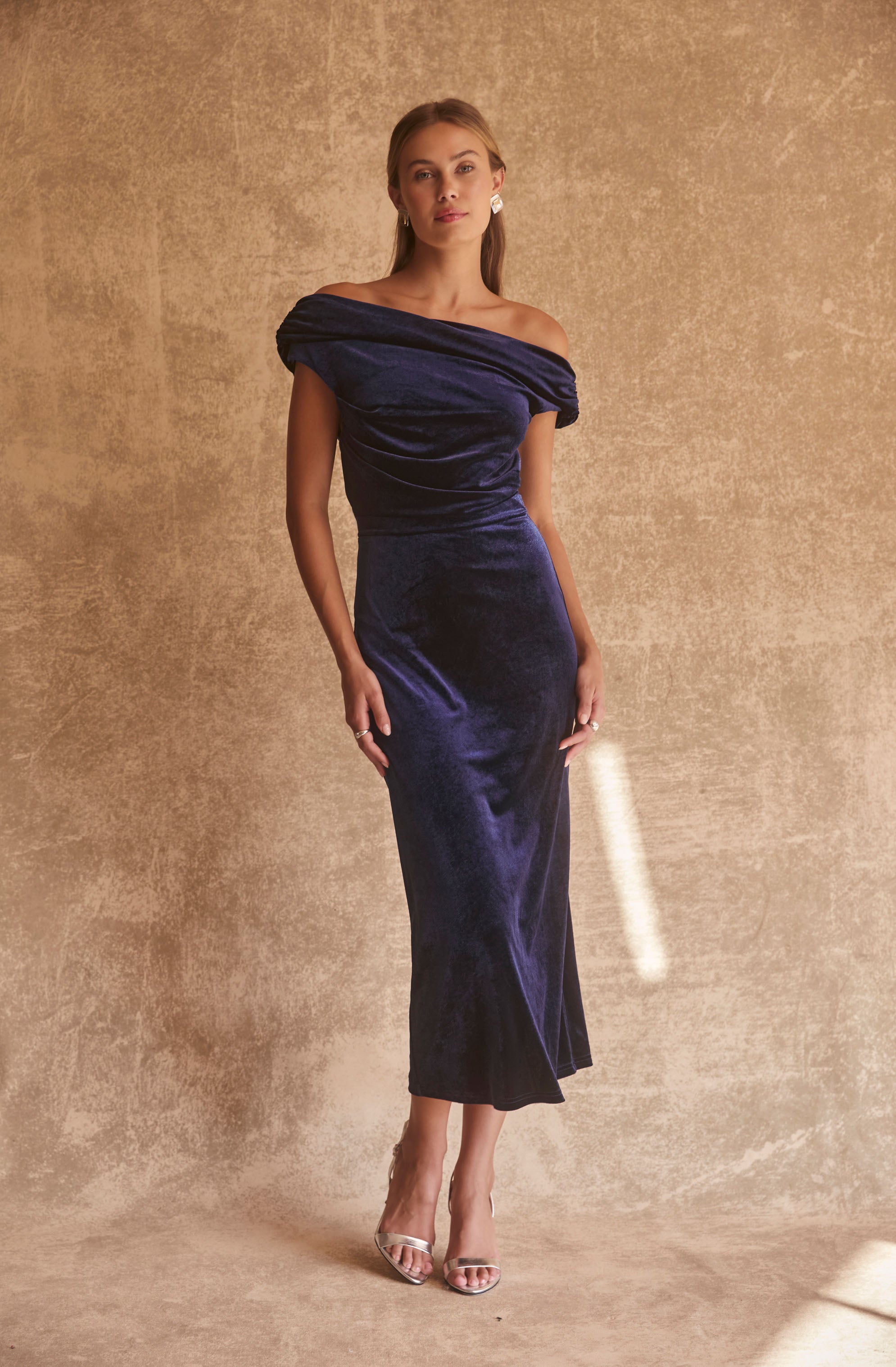 A woman is wearing an elegant, off-the-shoulder, deep blue velvet gown that is fitted and floor-length, with a draped neckline and a slit at the front. She is accessorized with statement earrings and is wearing clear high-heeled shoes.