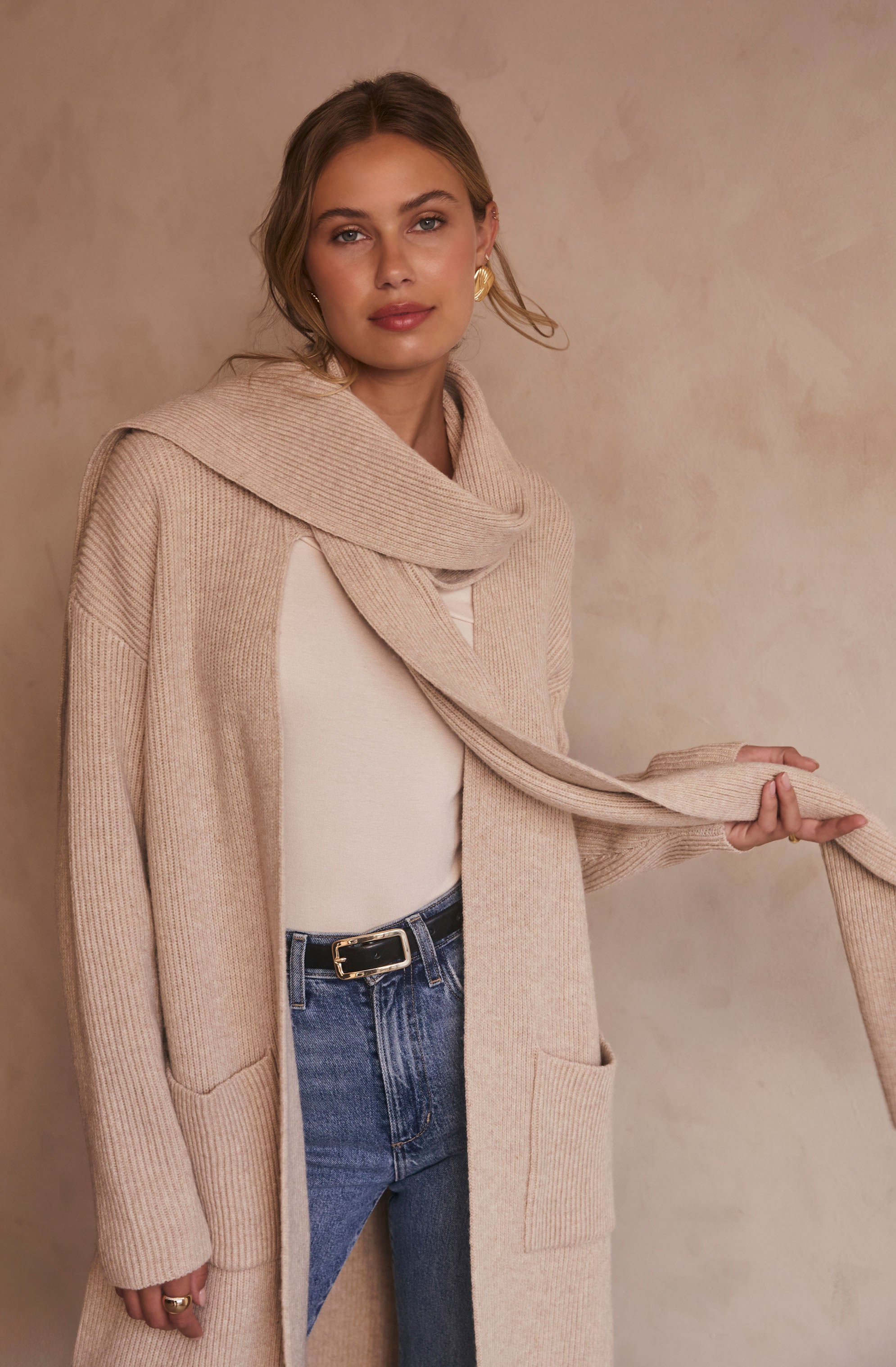 A woman is wearing a long beige knit cardigan with ribbed cuffs and large front pockets. Underneath, she has on a white top. She is also wearing medium-wash high-waisted jeans with a black belt that has a gold buckle. She has gold hoop earrings and a ring on her finger. The cardigan has a wide, draped collar that she is holding with one hand.