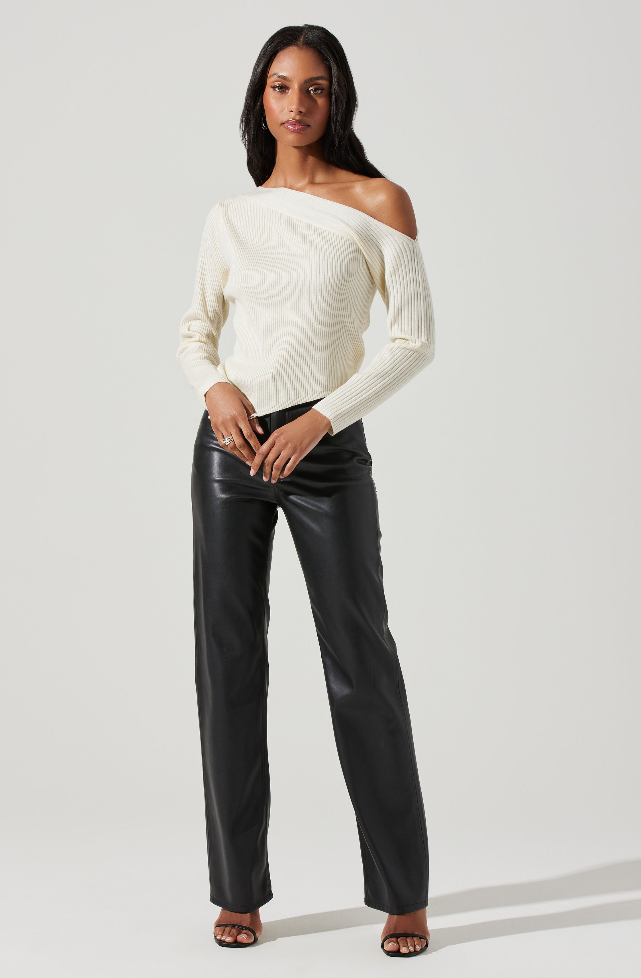 A woman is wearing a cream-colored, off-the-shoulder, ribbed knit sweater with long sleeves. She pairs it with high-waisted black leather pants that have a straight-leg fit. The outfit is completed with open-toe high heels.