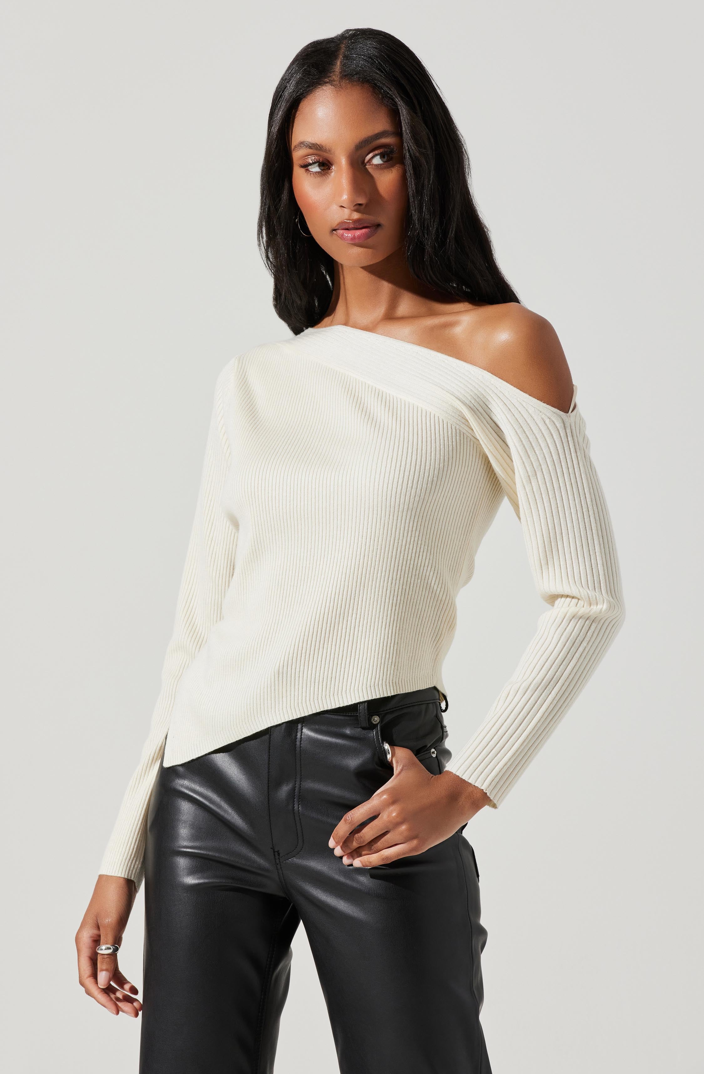 A woman is wearing a cream-colored, off-the-shoulder ribbed sweater with long sleeves. She has paired it with high-waisted black leather pants.