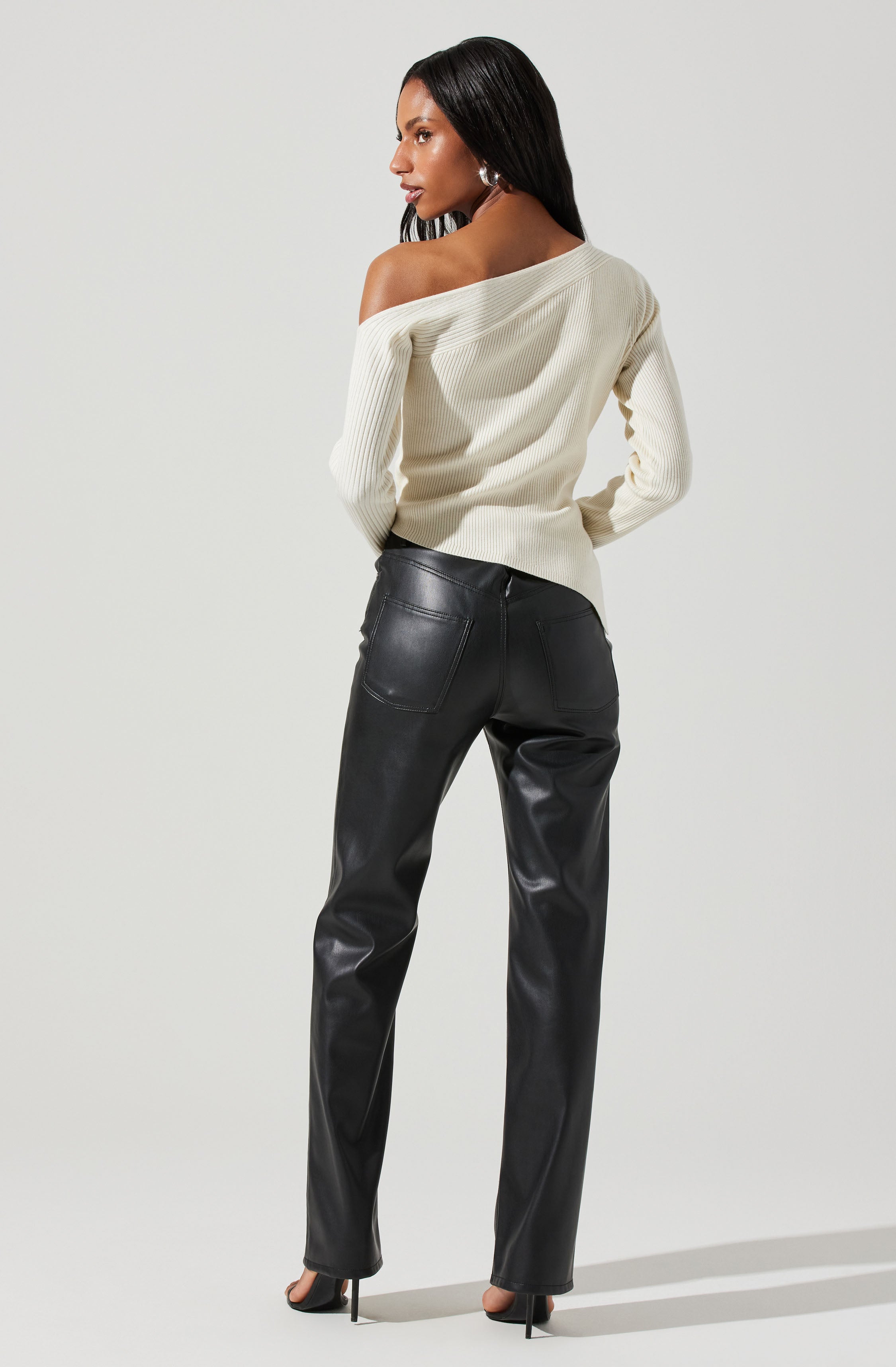 A woman is wearing an off-the-shoulder, long-sleeve, cream-colored ribbed sweater tucked into high-waisted, black leather pants with back pockets. She is also wearing high-heeled shoes and has long, straight, dark hair.