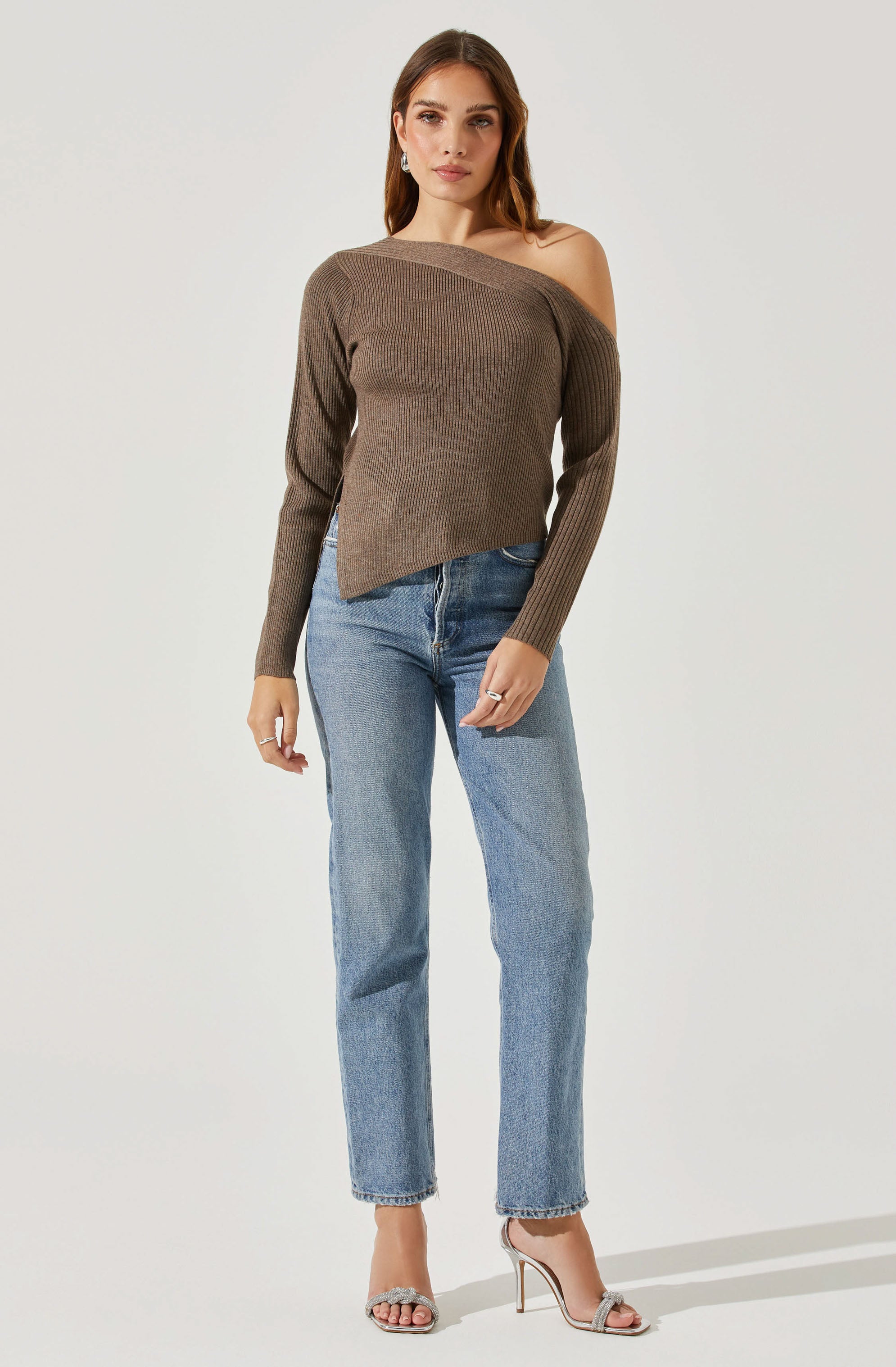 A woman is wearing an asymmetrical, off-the-shoulder, taupe-colored knit sweater with long sleeves and a ribbed texture. She is also dressed in light blue, high-waisted, straight-leg jeans. Her footwear consists of beige, open-toe high-heeled sandals with thin straps.