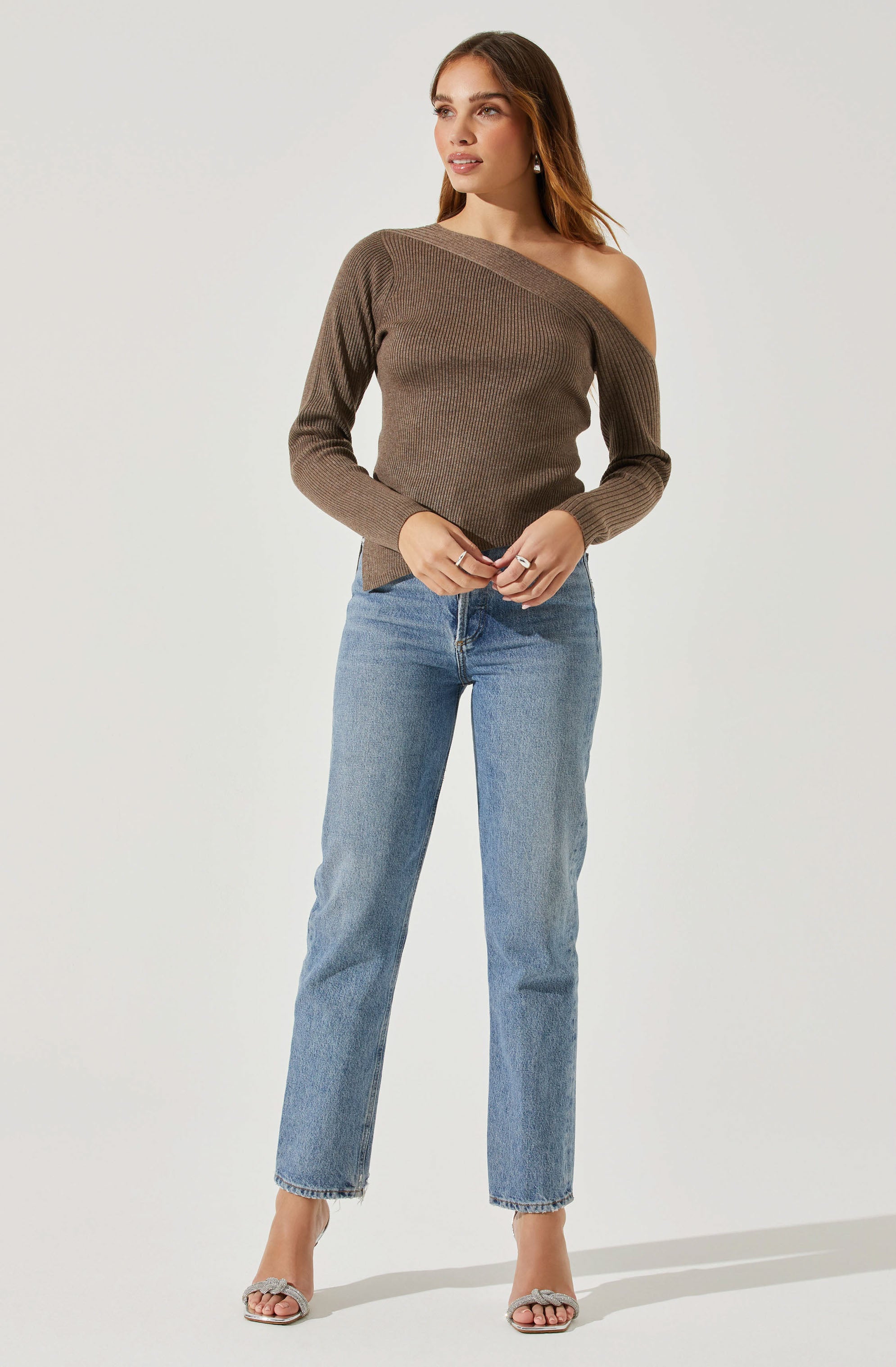 A woman is wearing a brown, ribbed, off-the-shoulder sweater with long sleeves. She is also wearing light blue, straight-leg jeans and open-toe heels with a chain detail.