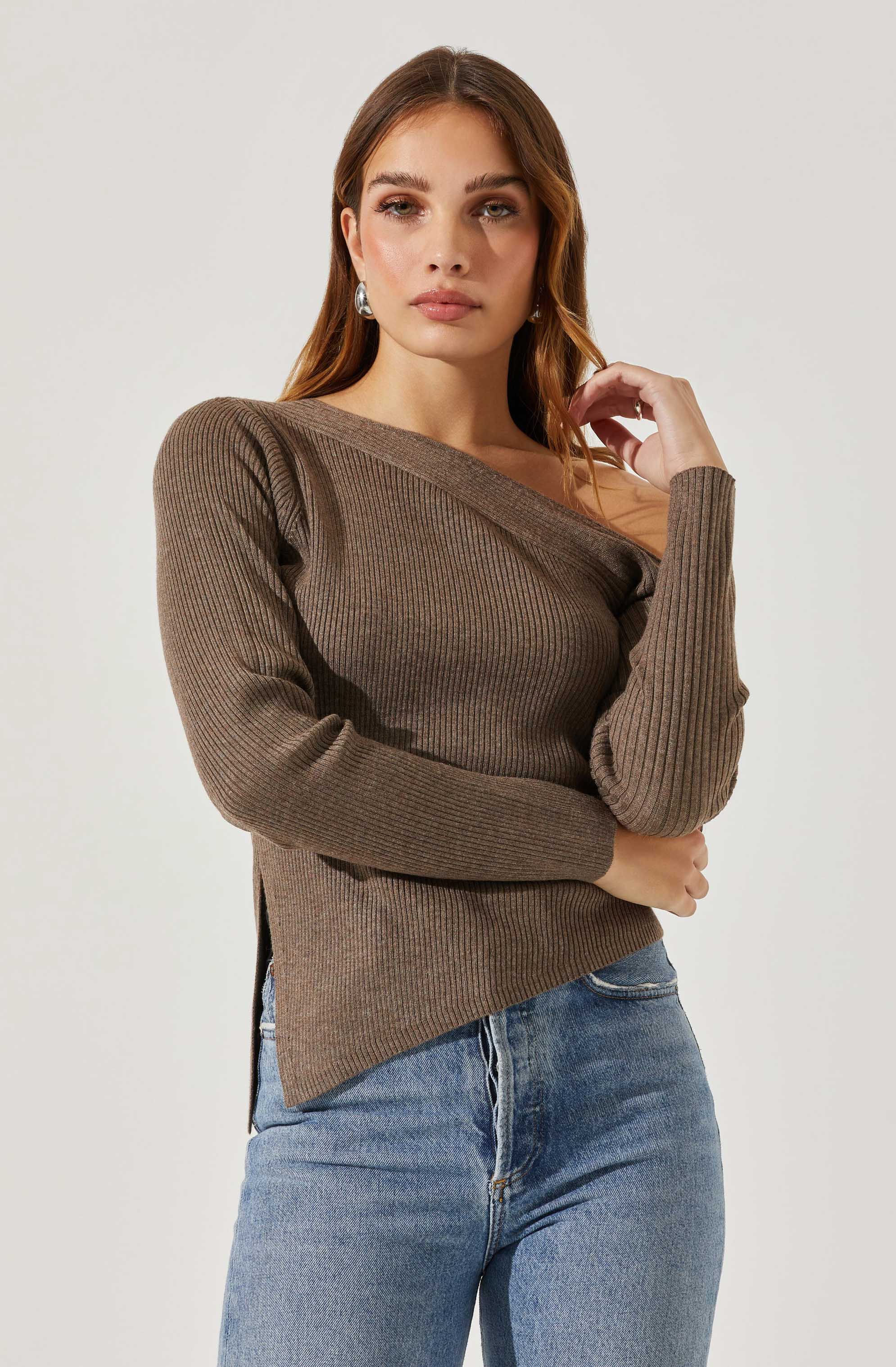 A woman is wearing a brown, ribbed, long-sleeve sweater with a wide neckline that reveals one shoulder. She pairs it with light blue jeans.