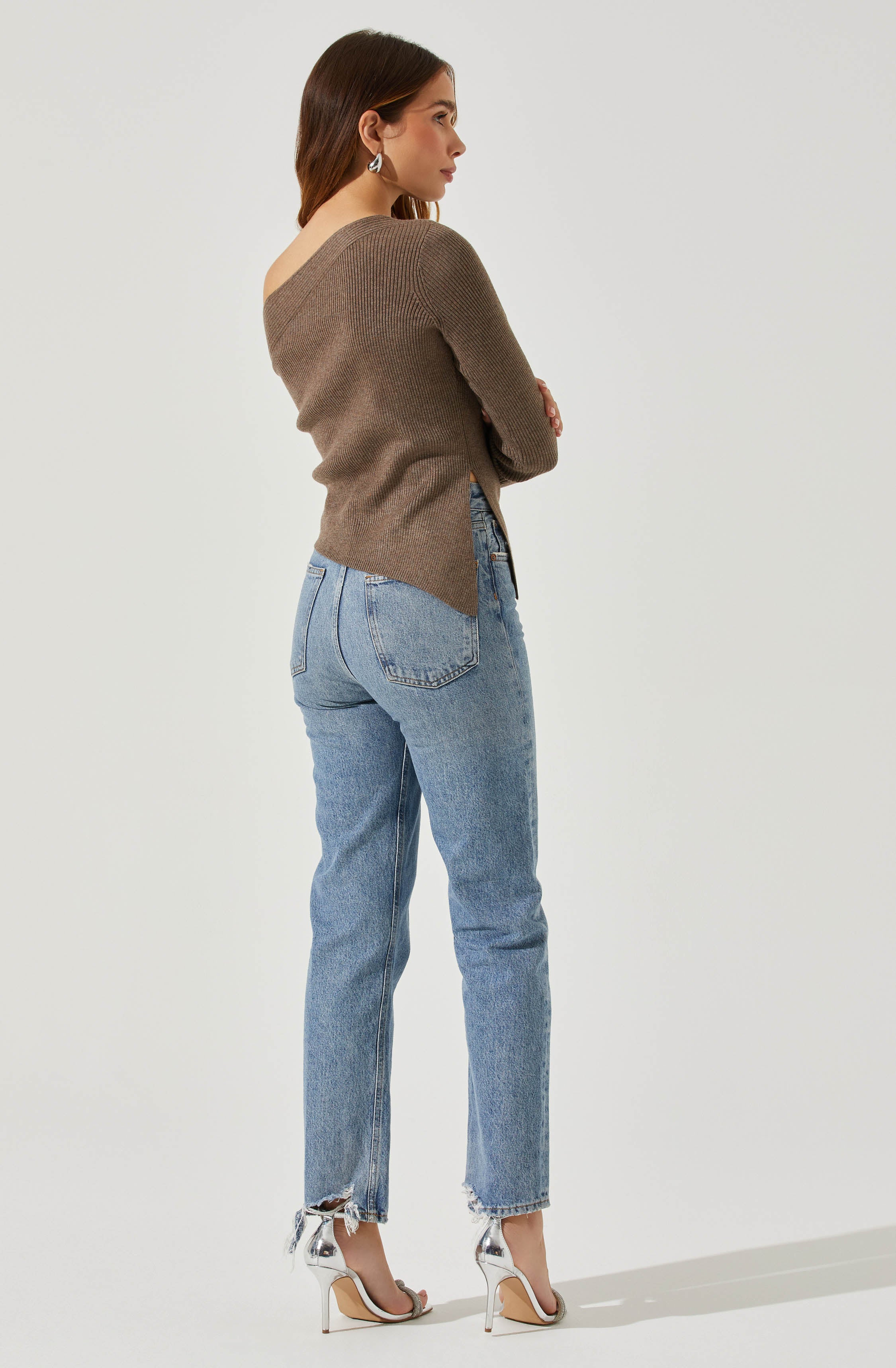 A woman is wearing a brown knitted sweater with one shoulder exposed. She is also wearing light blue high-waisted jeans with frayed hems at the ankles. She has on white high-heeled sandals with ankle straps and is accessorized with dangling earrings.