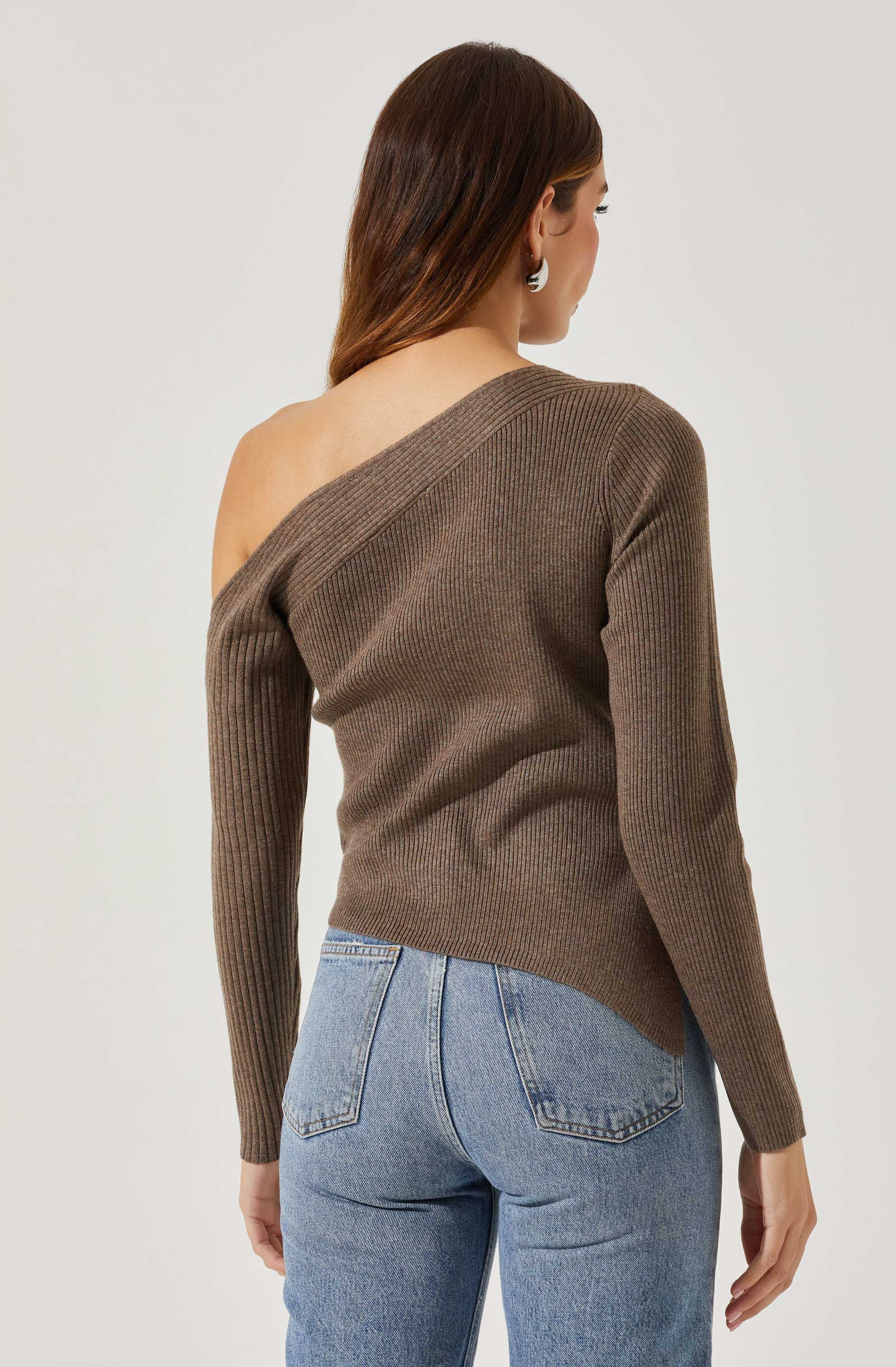 A woman is wearing a brown, ribbed, off-the-shoulder sweater with a diagonal neckline, paired with light blue high-waisted jeans. The sweater has long sleeves and a fitted silhouette.