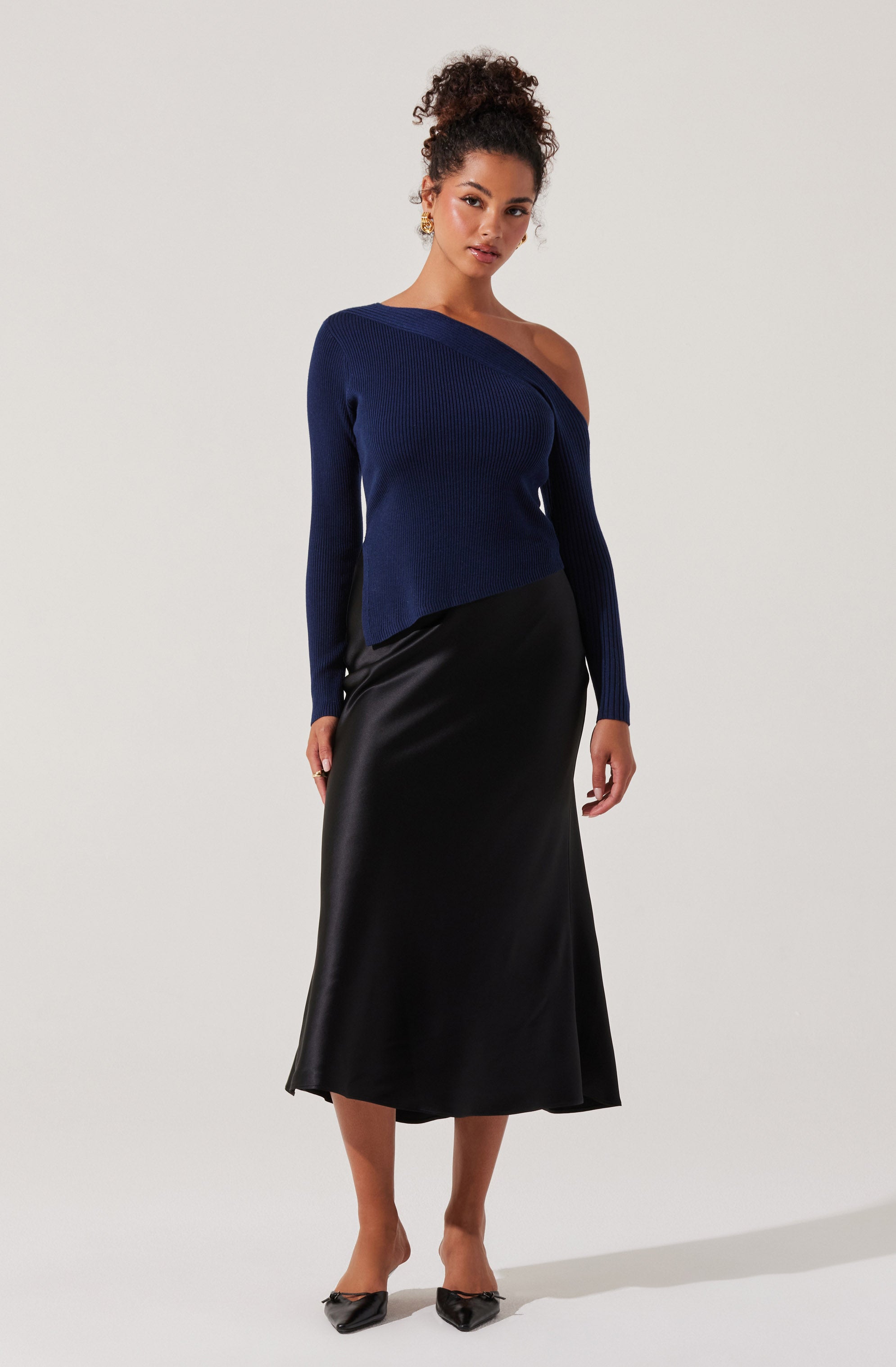 A woman is wearing a dark navy blue, asymmetrical, off-the-shoulder knitted sweater with a ribbed texture and a longer sleeve on one side. She pairs it with a black, midi-length, shiny satin slip skirt. She is also wearing black pointed-toe heels and gold earrings.