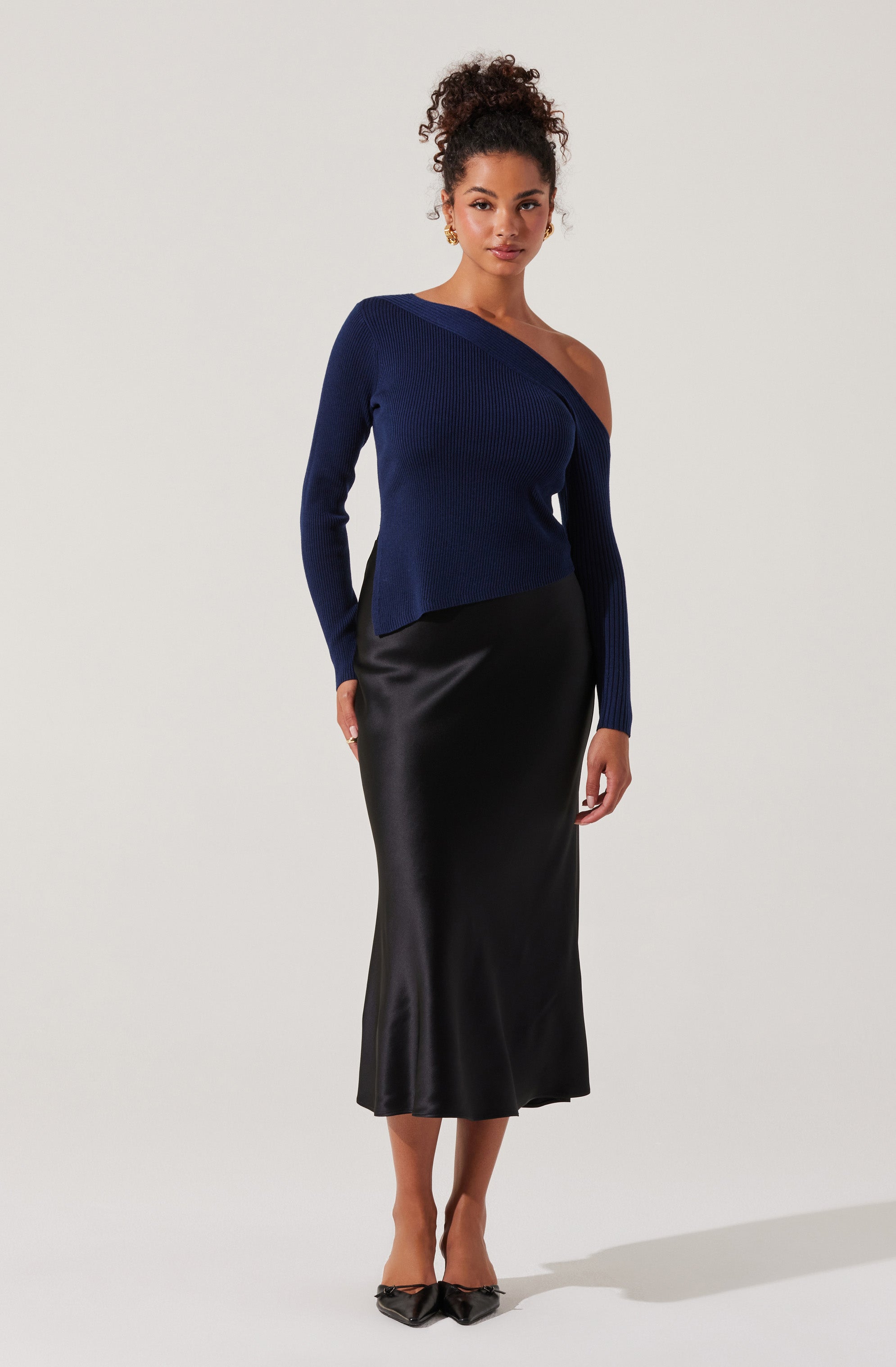 A woman is wearing a dark blue, asymmetrical, off-the-shoulder knit sweater with long sleeves, paired with a high-waisted, black satin, calf-length skirt. She completes her outfit with black pointed-toe heels.