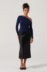 A woman is wearing a dark blue, asymmetrical, off-the-shoulder knit sweater with long sleeves, paired with a high-waisted, black satin, calf-length skirt. She completes her outfit with black pointed-toe heels.