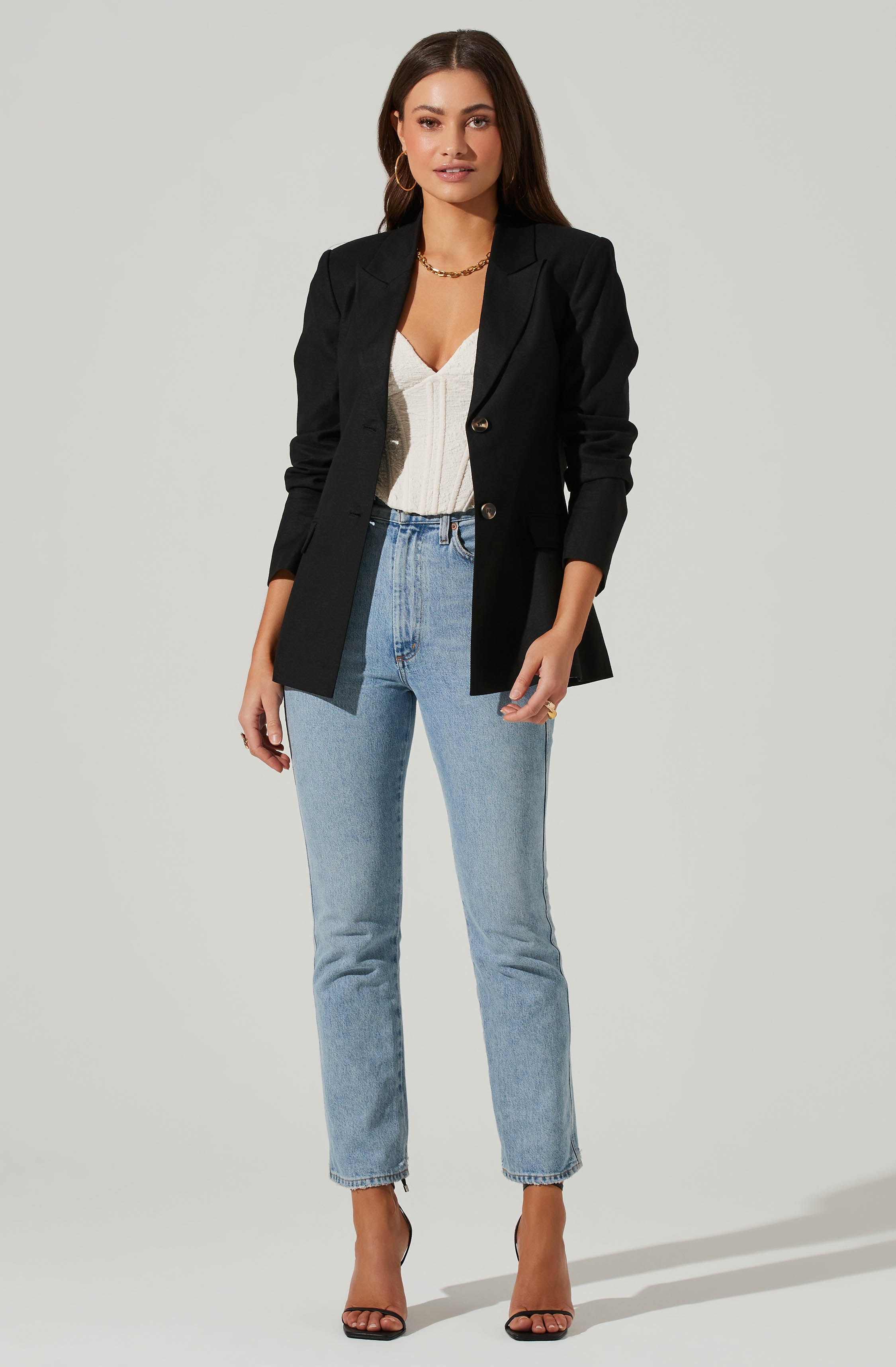A woman is wearing a fitted white top with a textured pattern, paired with light-wash high-waisted jeans. She has on a tailored black blazer with rolled-up sleeves, featuring gold buttons. She accessorizes with gold hoop earrings, a gold chain necklace, and rings, and completes the outfit with black high-heeled sandals.
