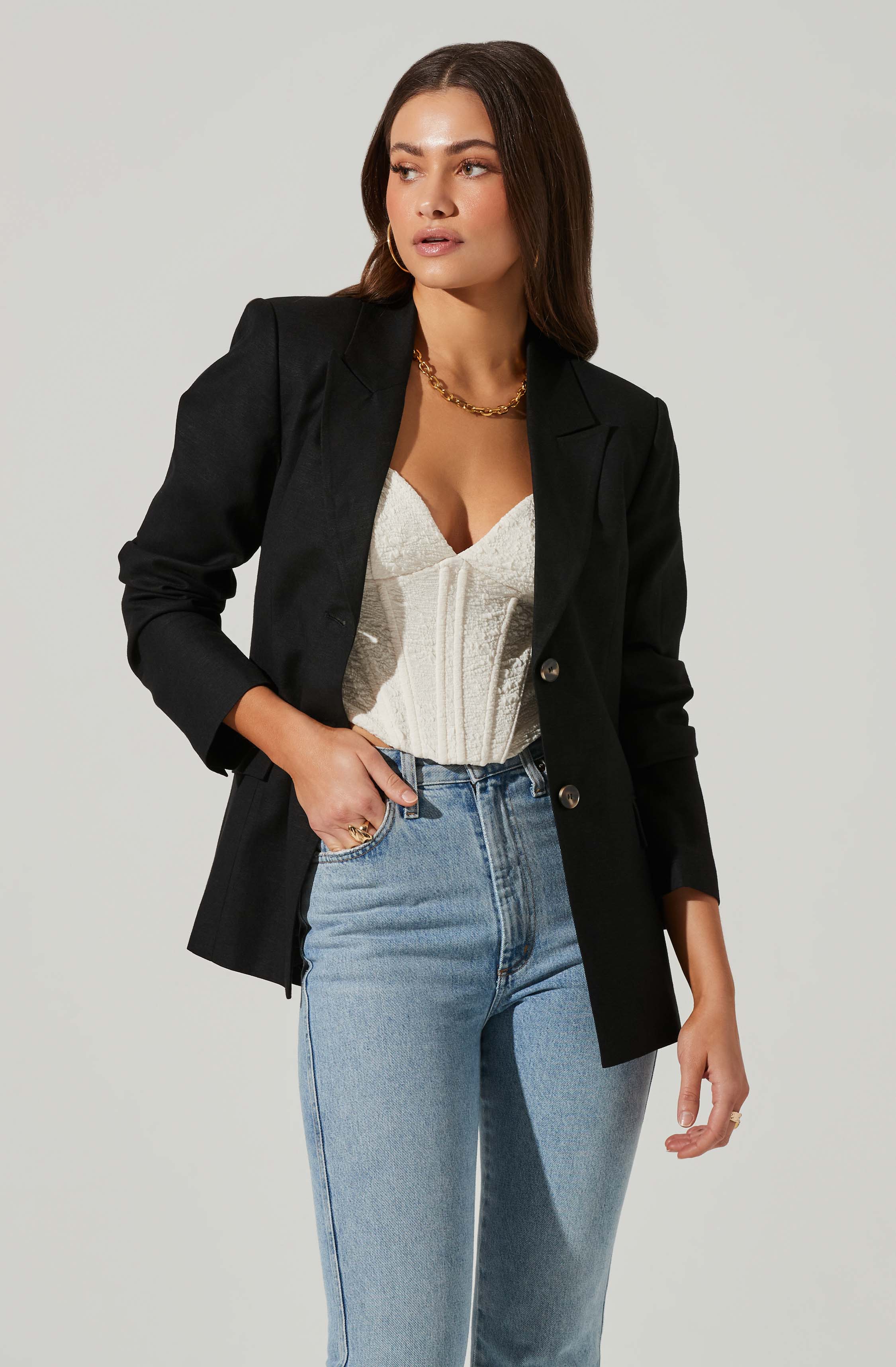 A woman is wearing a black blazer with rolled-up sleeves, a cream-colored textured corset top, and light blue high-waisted jeans. She accessorizes with a chunky gold necklace, hoop earrings, and rings on her fingers.