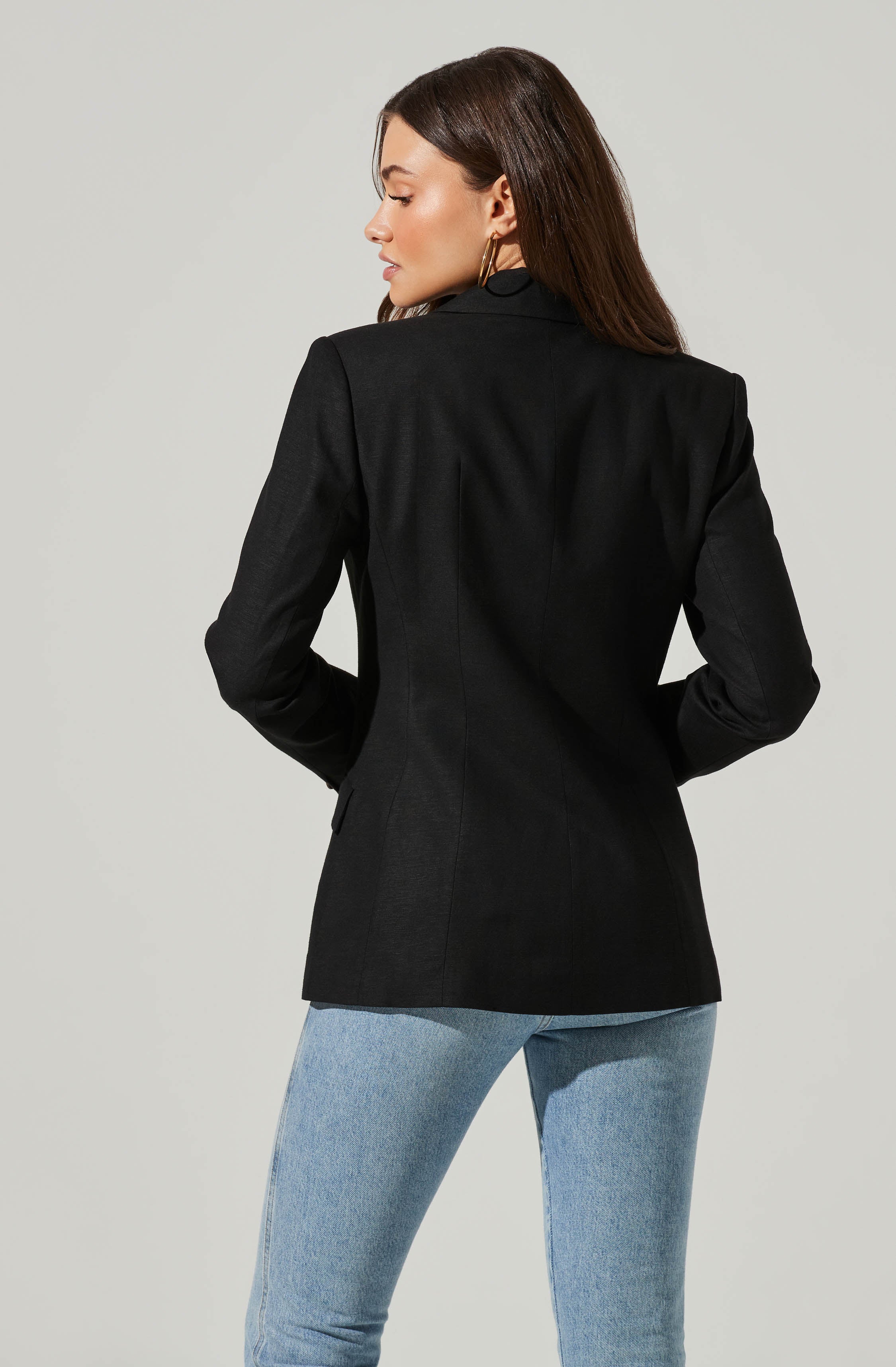 A woman is wearing a tailored black blazer with structured shoulders and a fitted waist. The blazer appears to have a smooth, slightly shiny texture. She is also wearing light blue high-waisted jeans. She has long, dark hair and is accessorized with large hoop earrings.