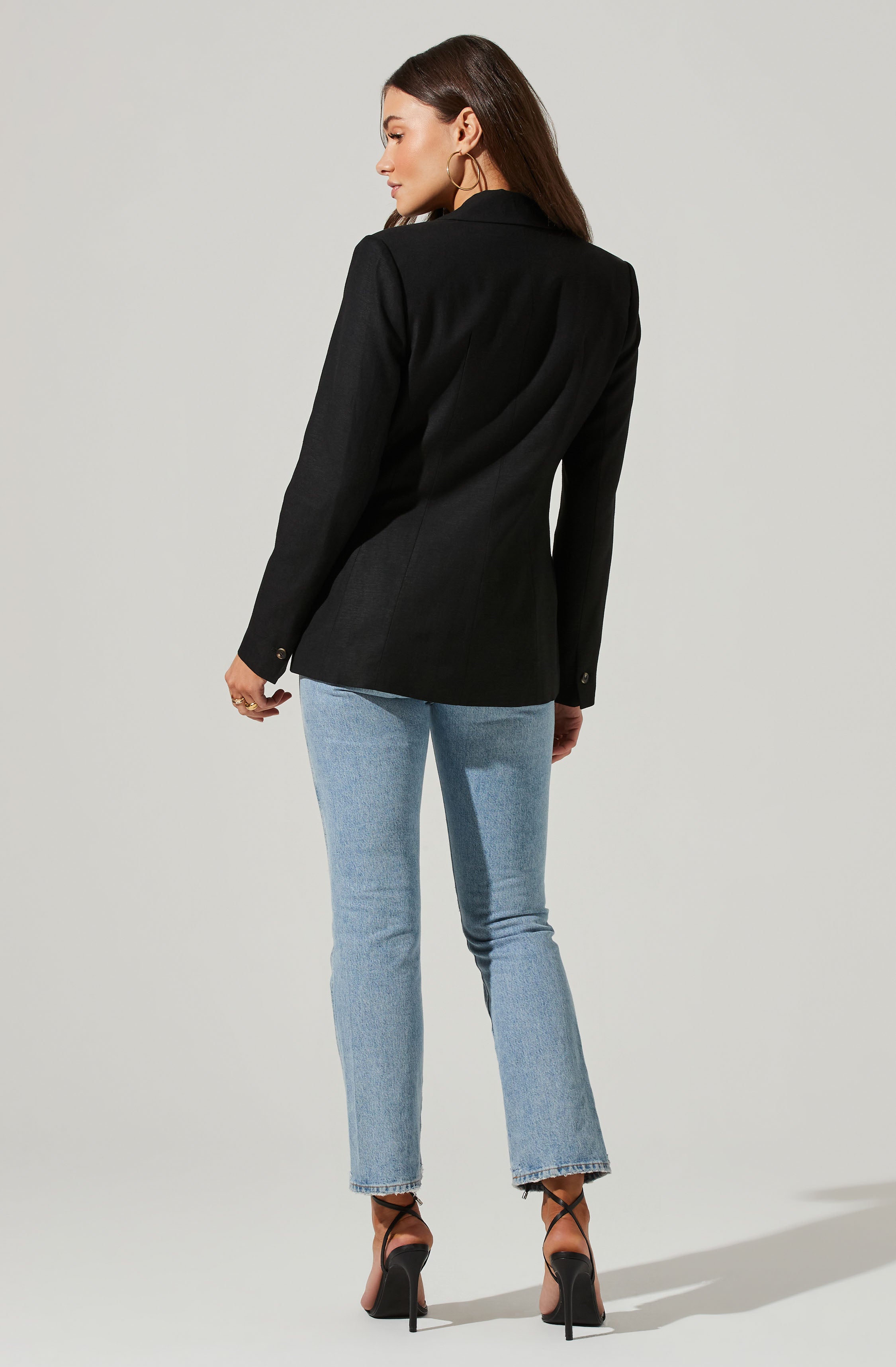A woman is wearing a tailored black blazer with a fitted silhouette, featuring a notched lapel, buttoned cuffs, and a smooth, structured fabric. She pairs it with light blue straight-leg jeans and black high-heeled sandals with thin ankle straps. She also wears gold hoop earrings.