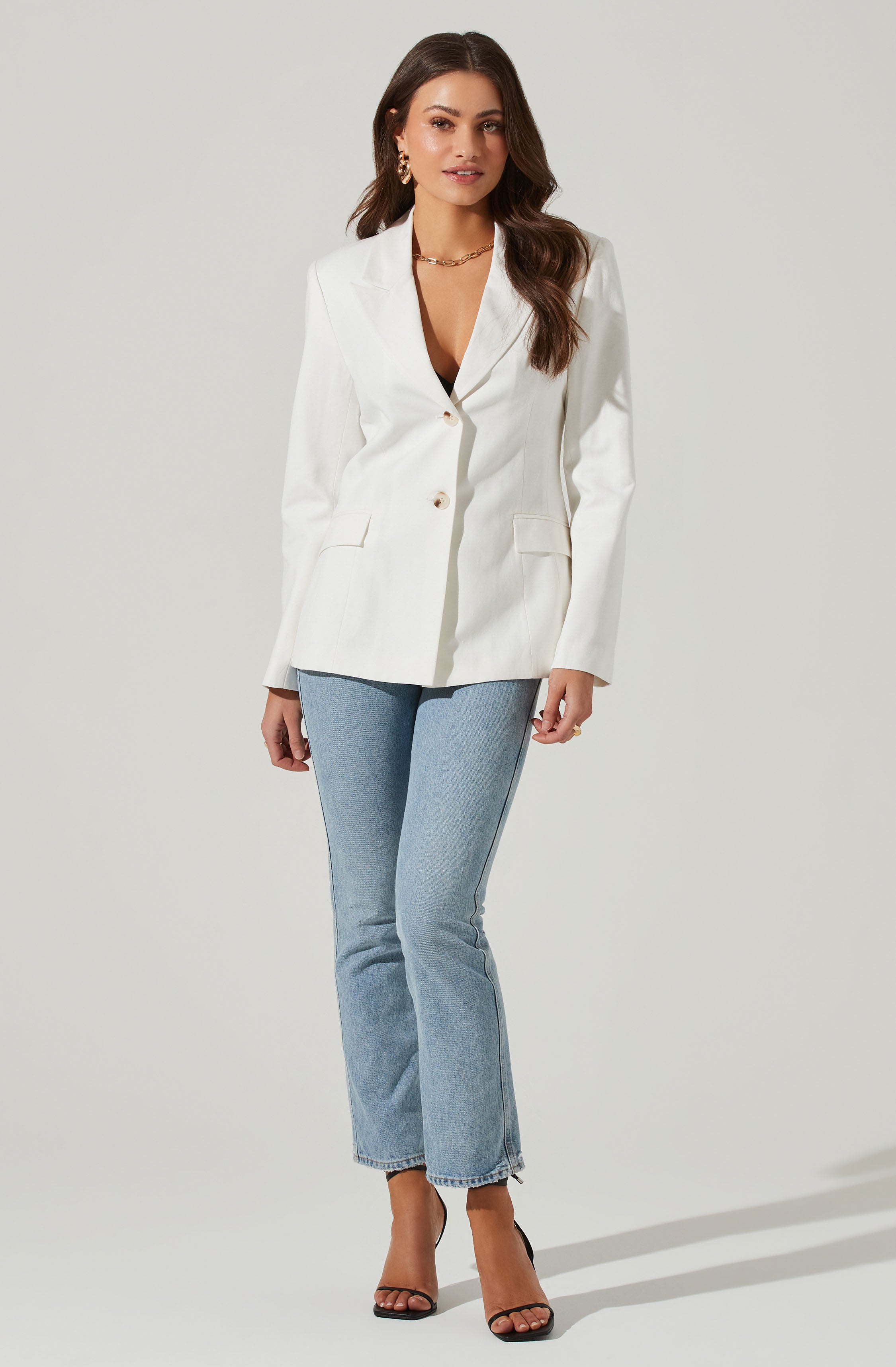 A woman is wearing a white blazer with a tailored fit, featuring a notched lapel collar and two large buttons down the front. She pairs it with light blue, high-waisted, straight-leg jeans that have a slightly frayed hem. She completes her outfit with black high-heeled sandals with ankle straps. She is accessorized with gold hoop earrings and a gold chain necklace.