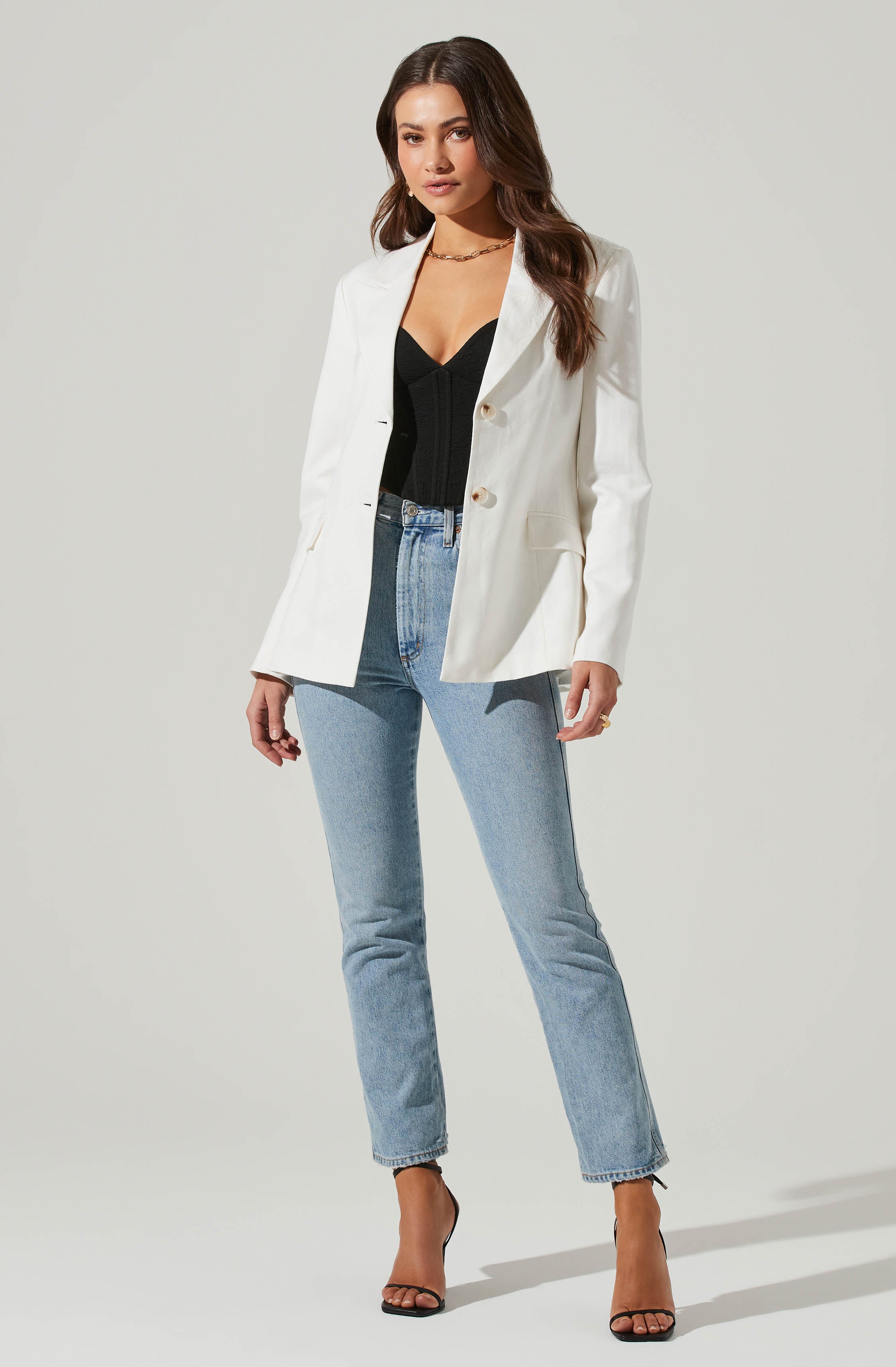 A woman is wearing a white blazer with large buttons and a tailored fit, paired with a black V-neck top. She has on light-wash, high-waisted jeans with a straight-leg cut, and is wearing black high-heeled sandals with thin straps. She accessorizes with a gold chain necklace and a gold ring on her right hand.