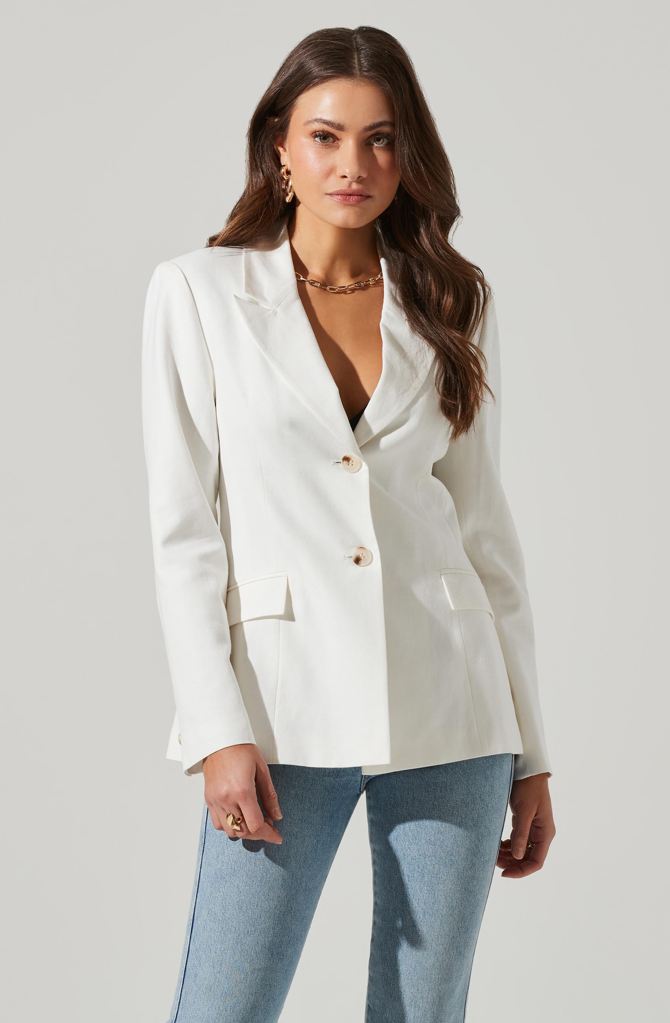 A woman is wearing a white blazer with a notched lapel collar, long sleeves, and two buttons down the front. The blazer features flap pockets at the waist. She is also wearing light blue jeans. She has accessorized with gold jewelry, including hoop earrings, a chain necklace, and rings.
