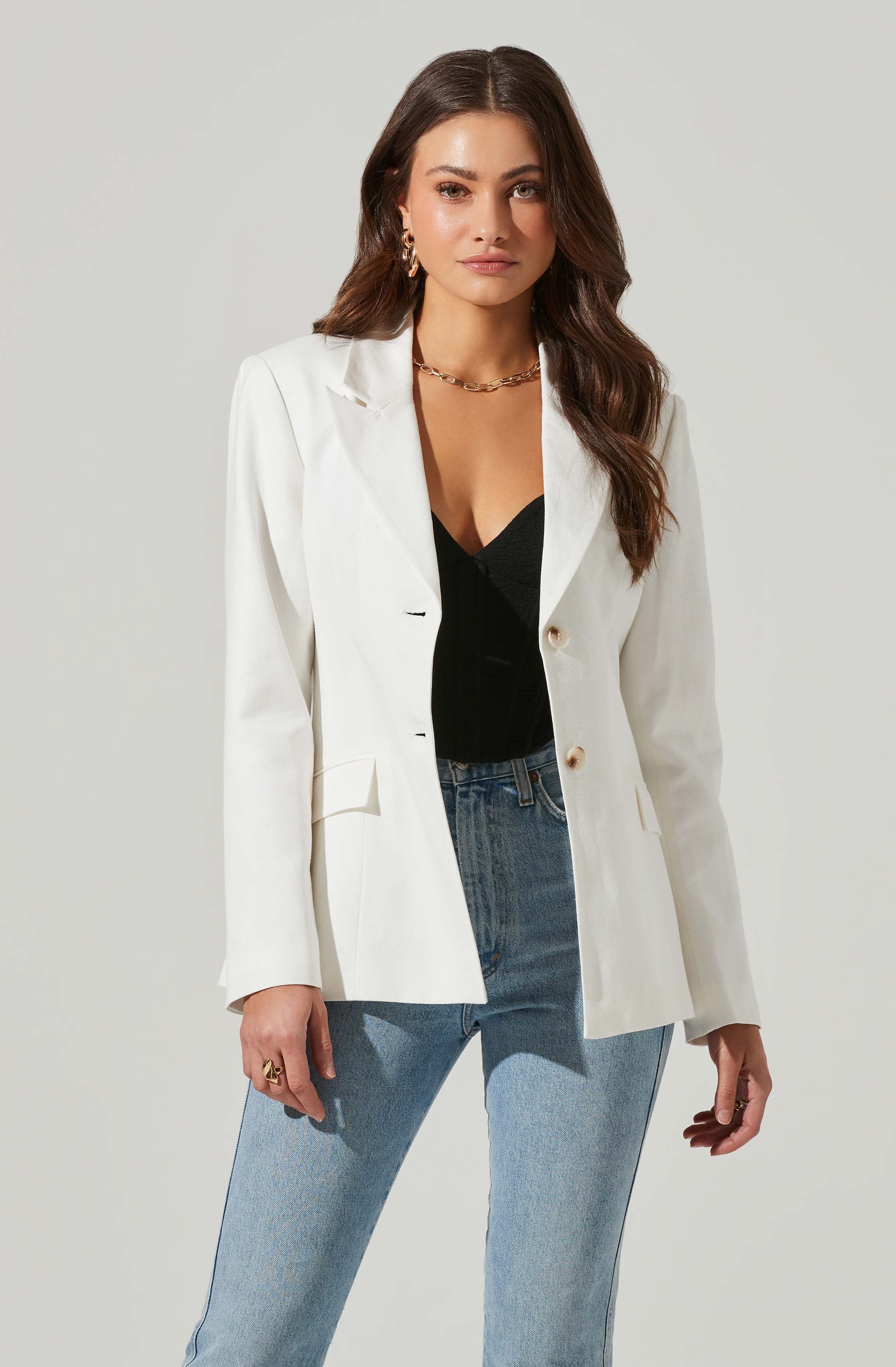 A woman is wearing a white blazer with a single button closure, a black top underneath, and light blue high-waisted jeans. She has earrings, a gold chain necklace, and a ring on her right hand.