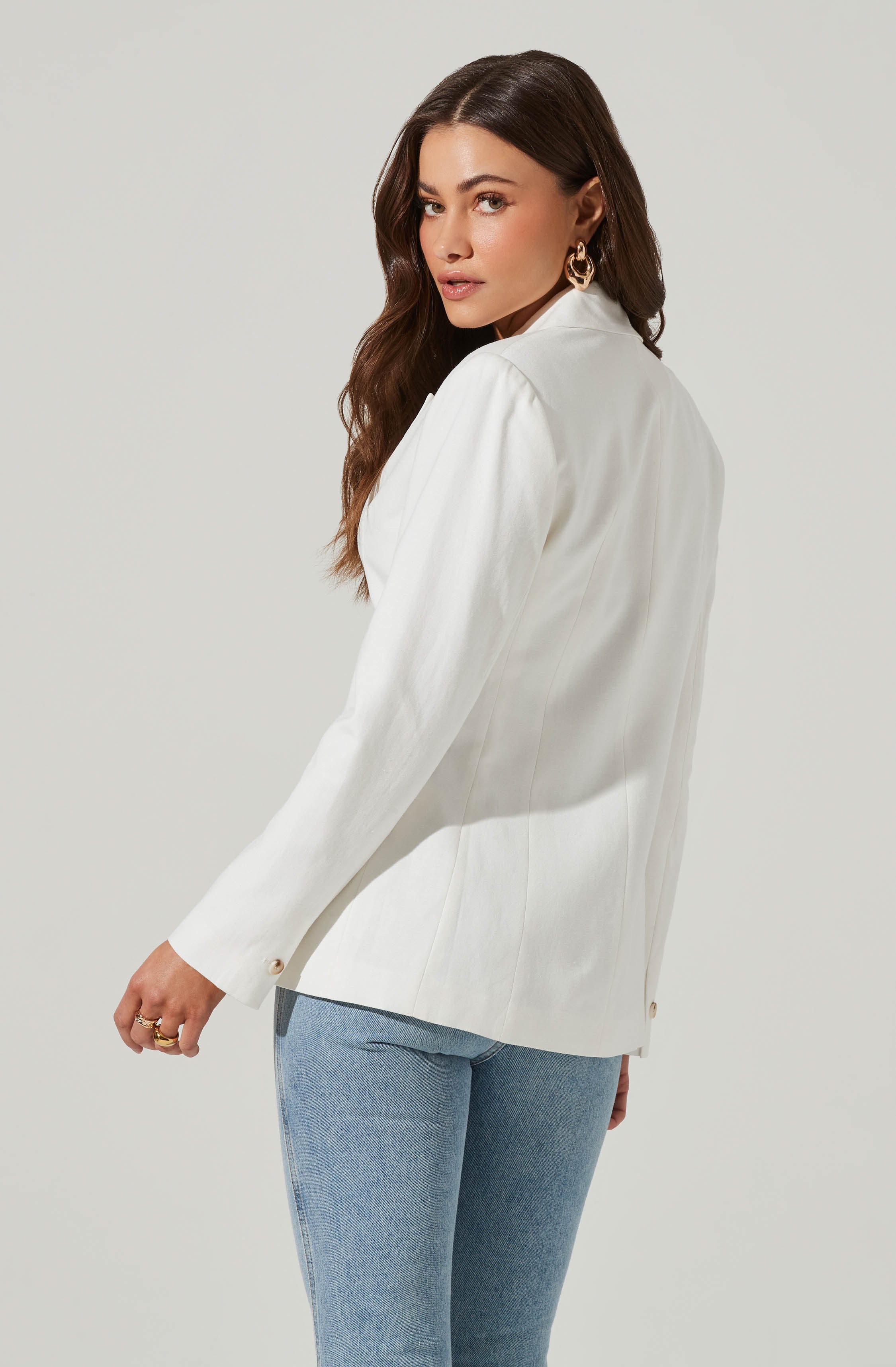 A woman is wearing a white blazer with long sleeves, featuring small, round, gold buttons at the cuffs. She has paired it with light blue denim jeans. The blazer appears tailored, with a smooth texture and a structured fit. Her accessories include large, gold, hoop earrings.