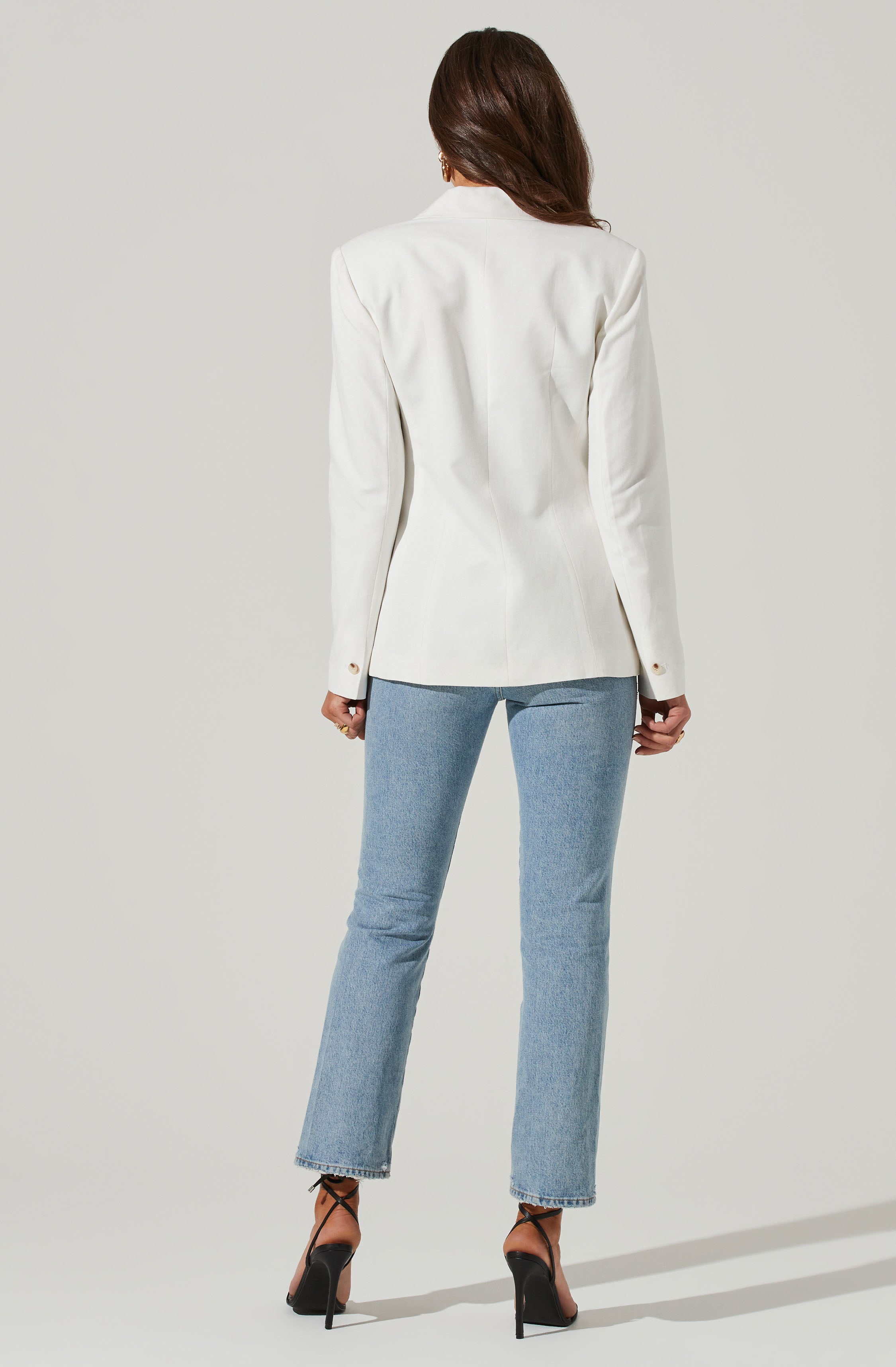 A woman is wearing a white blazer with structured shoulders and buttoned cuffs, paired with light blue jeans. She is also wearing black high-heeled shoes with ankle straps.