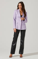 A woman is wearing a lavender blazer with a notched lapel, single button closure, and flap pockets. She has paired it with black leather or faux leather pants and black open-toe heels. She accessorizes with gold hoop earrings, a gold chain necklace, and gold rings.