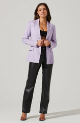 A woman is wearing a light purple blazer with a classic lapel collar and structured shoulders. She has on a black crop top underneath and black high-waisted pants with a slightly shiny leather or faux leather appearance. She accessorizes with gold hoop earrings, a gold chain necklace, and gold rings on her fingers. She is also wearing black high-heeled sandals with thin straps.