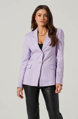 A woman is wearing a lavender blazer with structured shoulders, two buttons, and flap pockets. She has paired it with black pants that appear to be made of a shiny material, possibly leather or faux leather. She accessorizes with gold hoop earrings, a gold chain necklace, and a gold ring. Underneath the blazer, she is wearing a black top.